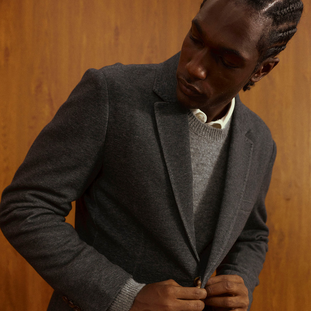 Man in a gray blazer over a gray crewneck sweater and white collared shirt, against a warm wood-paneled wall.