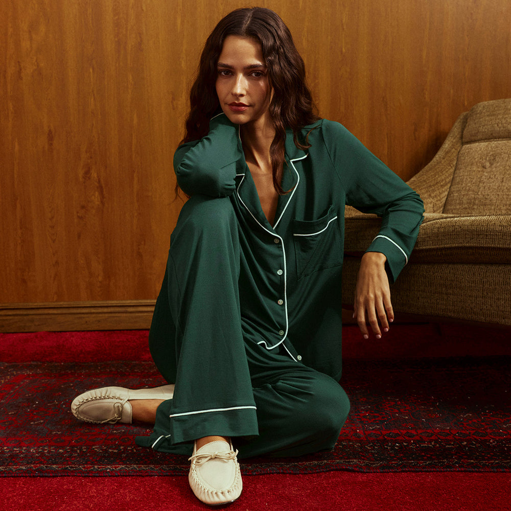 Woman seated on a red patterned rug in a green pajama set with white piping and cream moccasin flats, beside a tan armchair and wood-paneled wall.