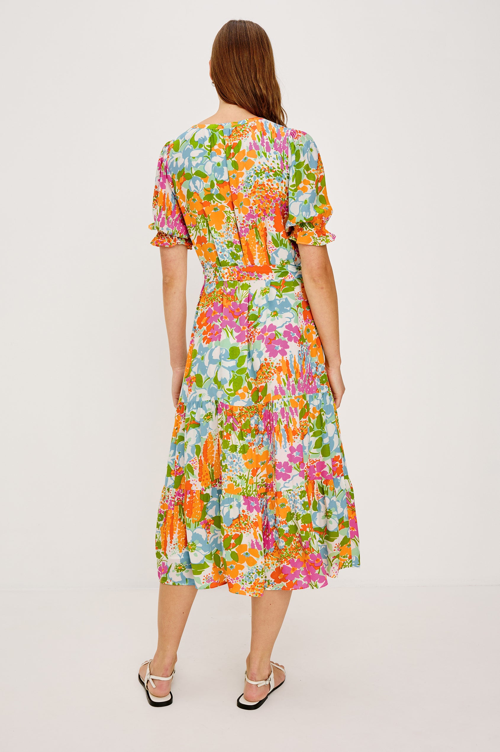 Eliana Midi Dress with Elbow-Length Sleeves with Shirred Cuffs, a V Neck, Buttons Down the Bodice, a Fabric Tie at the Waist  and a Tiered Skirt in Day Garden Colorway, a Floral Pattern in Blue, Pink and Oranges on White - Back View Featured on Model