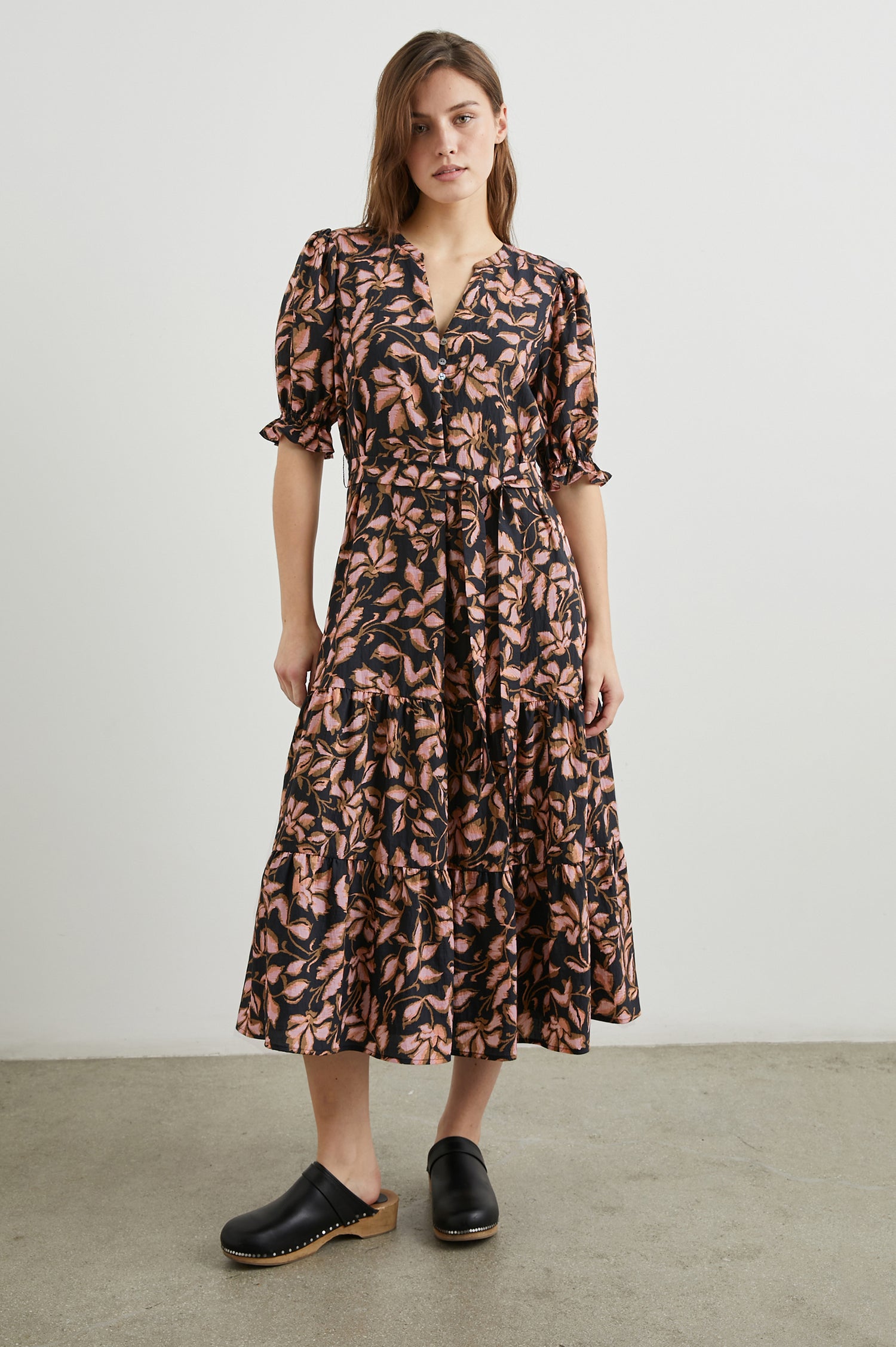 Eliana Midi Dress with Elbow-Length Sleeves with Shirred Cuffs, a V Neck, Buttons Down the Bodice, a Fabric Tie at the Waist  and a Tiered Skirt in Jasmine Colorway, a Floral Pattern in Pink and Brown on Black - Front Full Body View Featured on Model