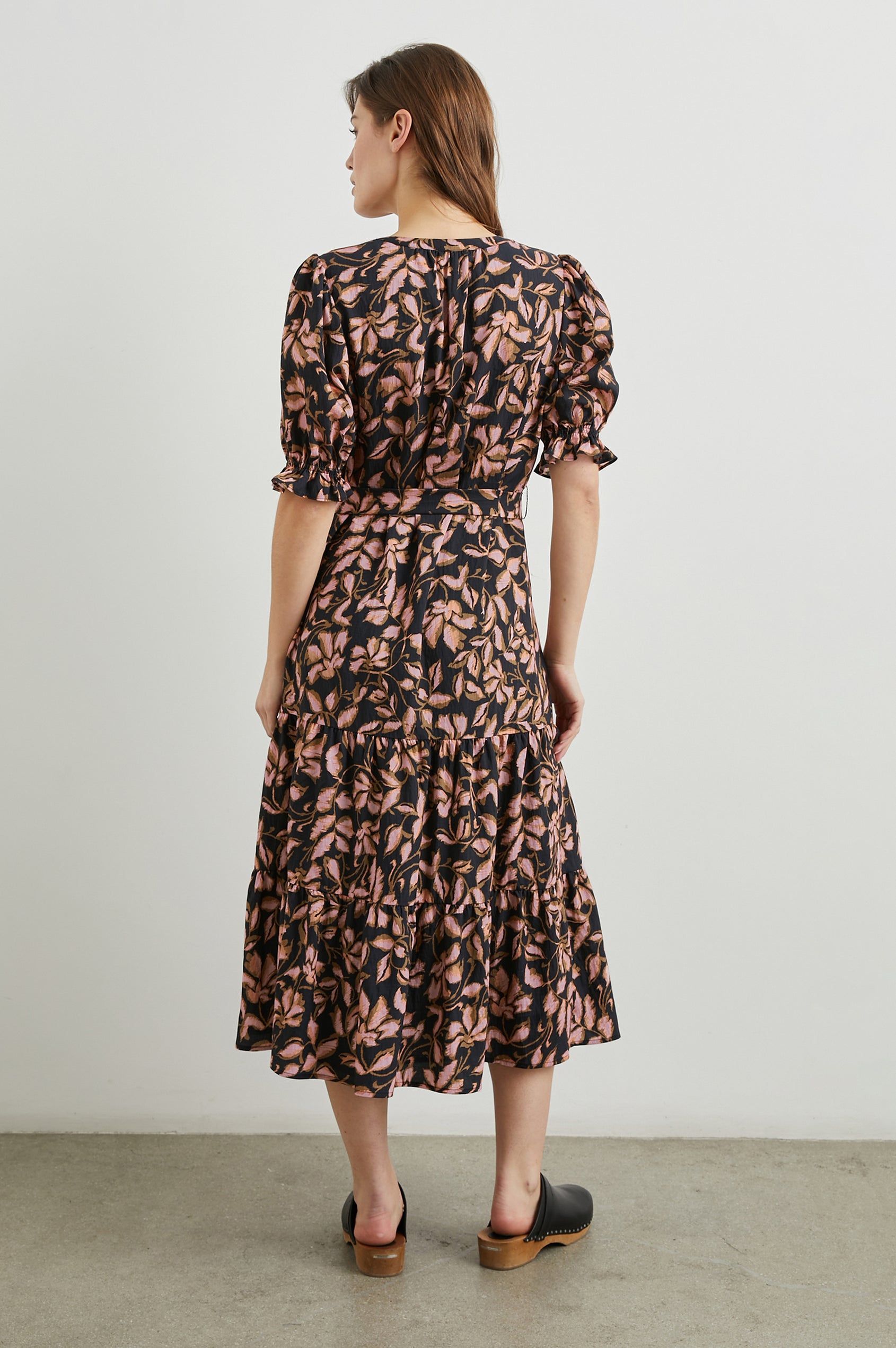 Eliana Midi Dress with Elbow-Length Sleeves with Shirred Cuffs, a V Neck, Buttons Down the Bodice, a Fabric Tie at the Waist  and a Tiered Skirt in Jasmine Colorway, a Floral Pattern in Pink and Brown on Black - Back Full Body View Featured on Model