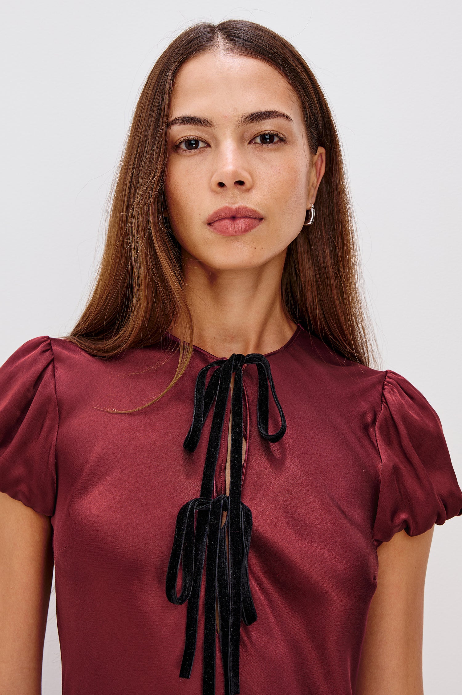 A garnet satin-back crepe midi dress cut on the bias. The dress features short puff sleeves, a deep V-neckline with velvet bow self-ties, and a fluid, flattering silhouette. Close Up View Featured on Model.