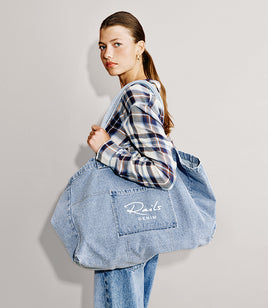 A woman wearing a blue plaid shirt and jeans carries a large light blue denim tote bag with a front pocket featuring the embroidered brand name 'Ravels DENIM'.
