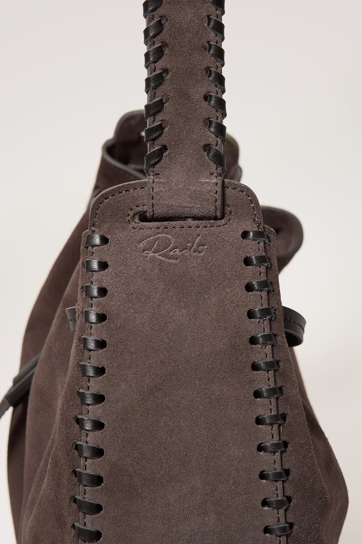 Firenze Bucket Bag in Suede With Whipstitch Trim, Drawcord Closure, and Interior Zip Pocket in Dark Brown Suede Colorway - close up side view of exterior, showing rails logo at the base of the strap.