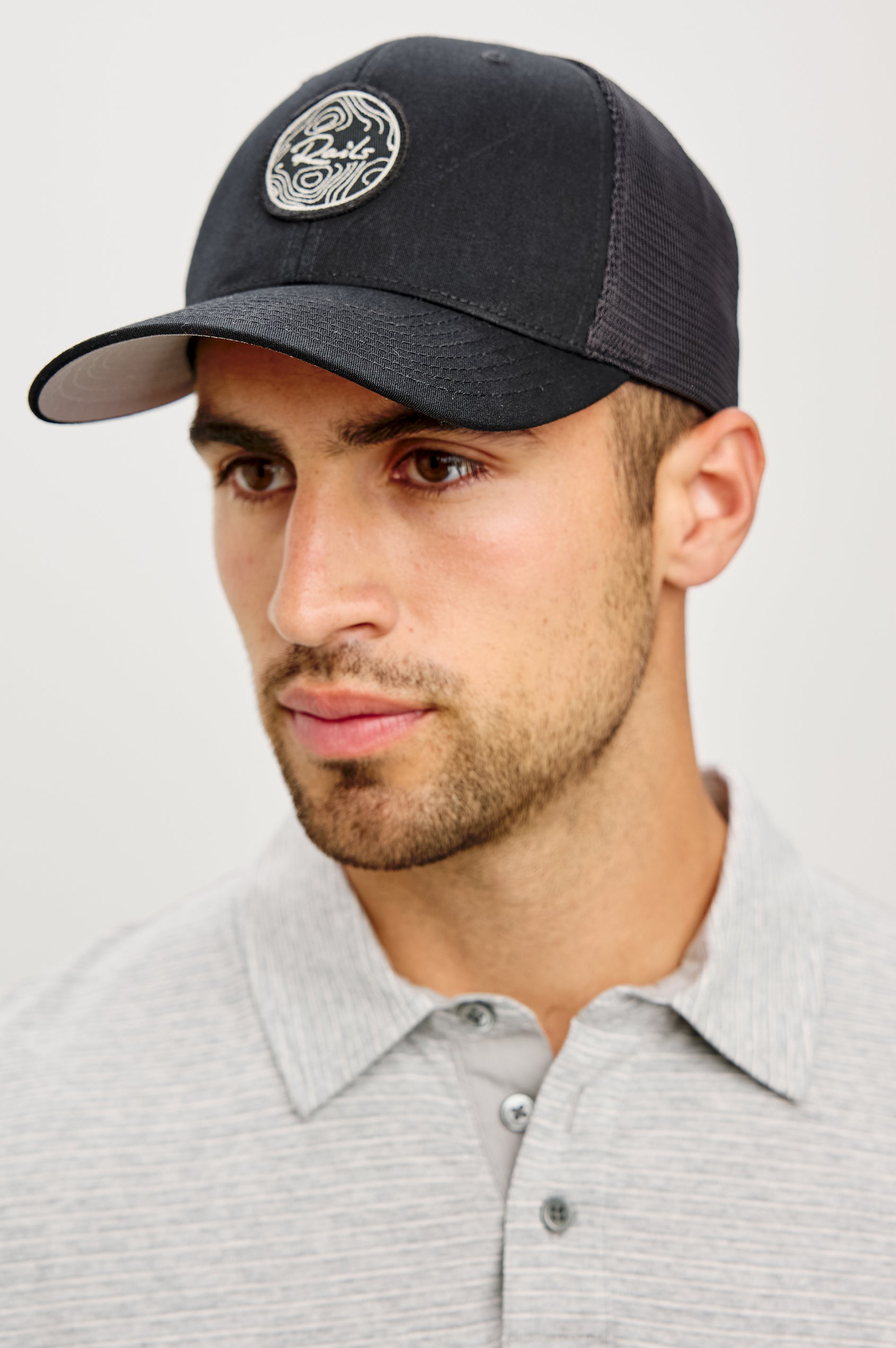 Golf Hat With Embroidered Branded Patch at Front, Adjustable Snapback, Mesh Back, and 6-Panel Construction in Black Colorway - Upper Body Front View Featured on Model