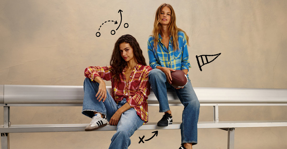 Two women sitting on a silver metal bleacher bench against a textured beige wall. The woman on the left wears a red and brown plaid shirt with blue jeans, and the woman on the right wears a blue and yellow plaid shirt with blue jeans.
