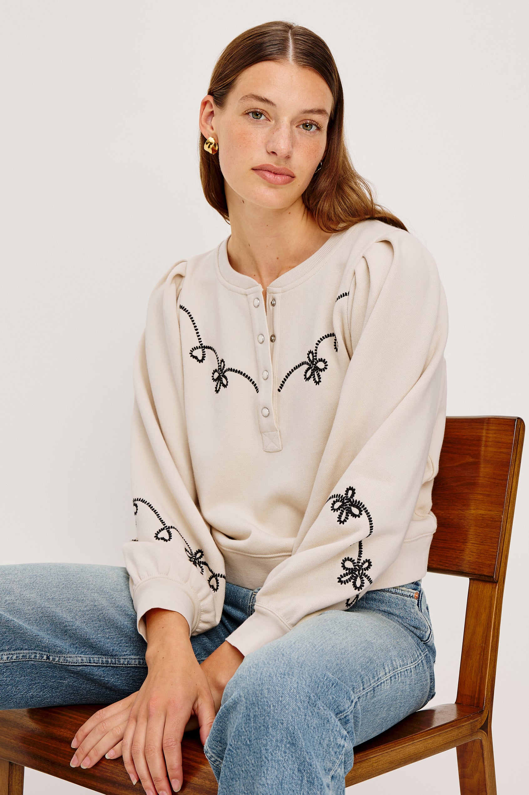 The Hanne is a henley style long sleeve sweatshirt with a snap front placket, rib trim, western style contrast black embroidery and puff sleeves in Ecru colorway. Front seated view featured on model.