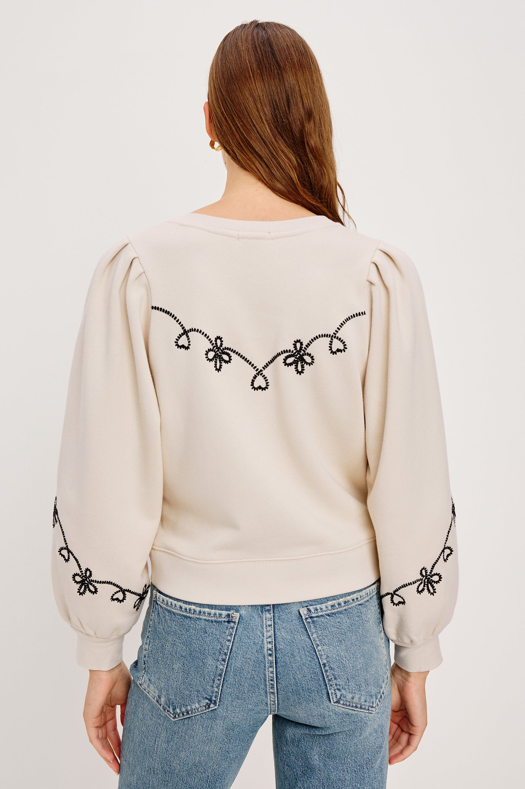 The Hanne is a henley style long sleeve sweatshirt with a snap front placket, rib trim, western style contrast black embroidery and puff sleeves in Ecru colorway. Back seated view featured on model.