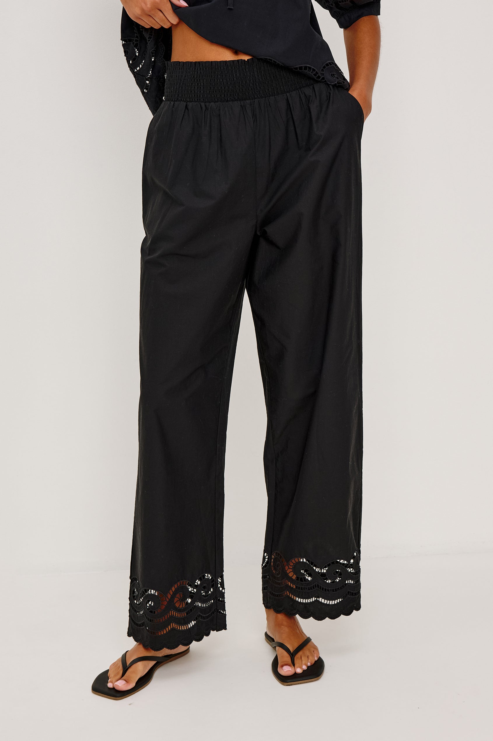 Henrik High-Rise High-Waisted Pant With Wide Smocked Waistband, Side Pockets, Straight Legs, and Scalloped Ladder Embroidered Hem in Black Colorway - Lower Body Front View Featured on Model