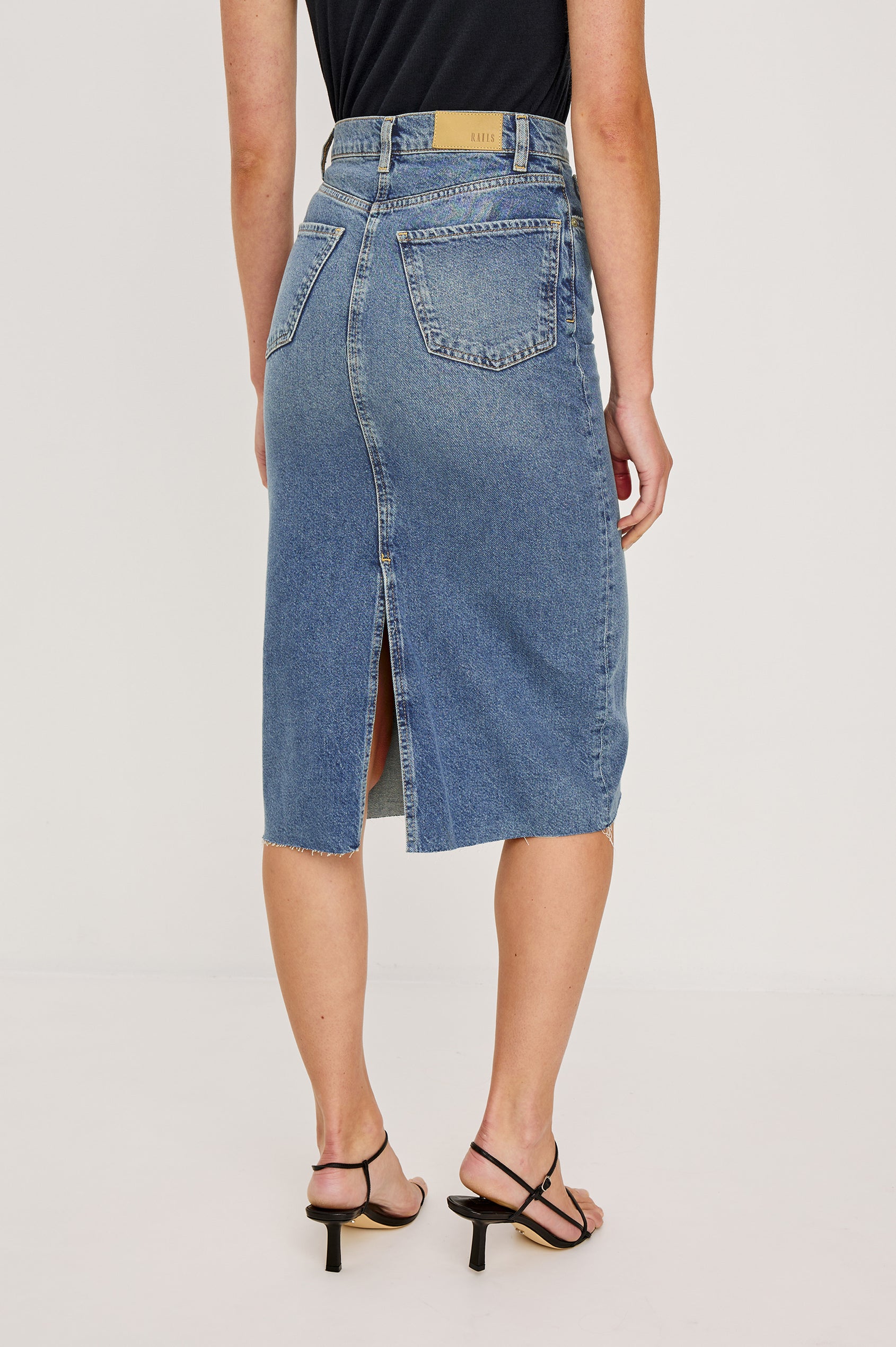 Highland Denim Midi Skirt with a Raw Hem, a Back Vent, Additional Front Seams, Belt Loops, Two Front Pockets plus a Coin Pocket and Two Back Patch Pockets in Vintage Sapphire Colorway - Back View Featured on Model 