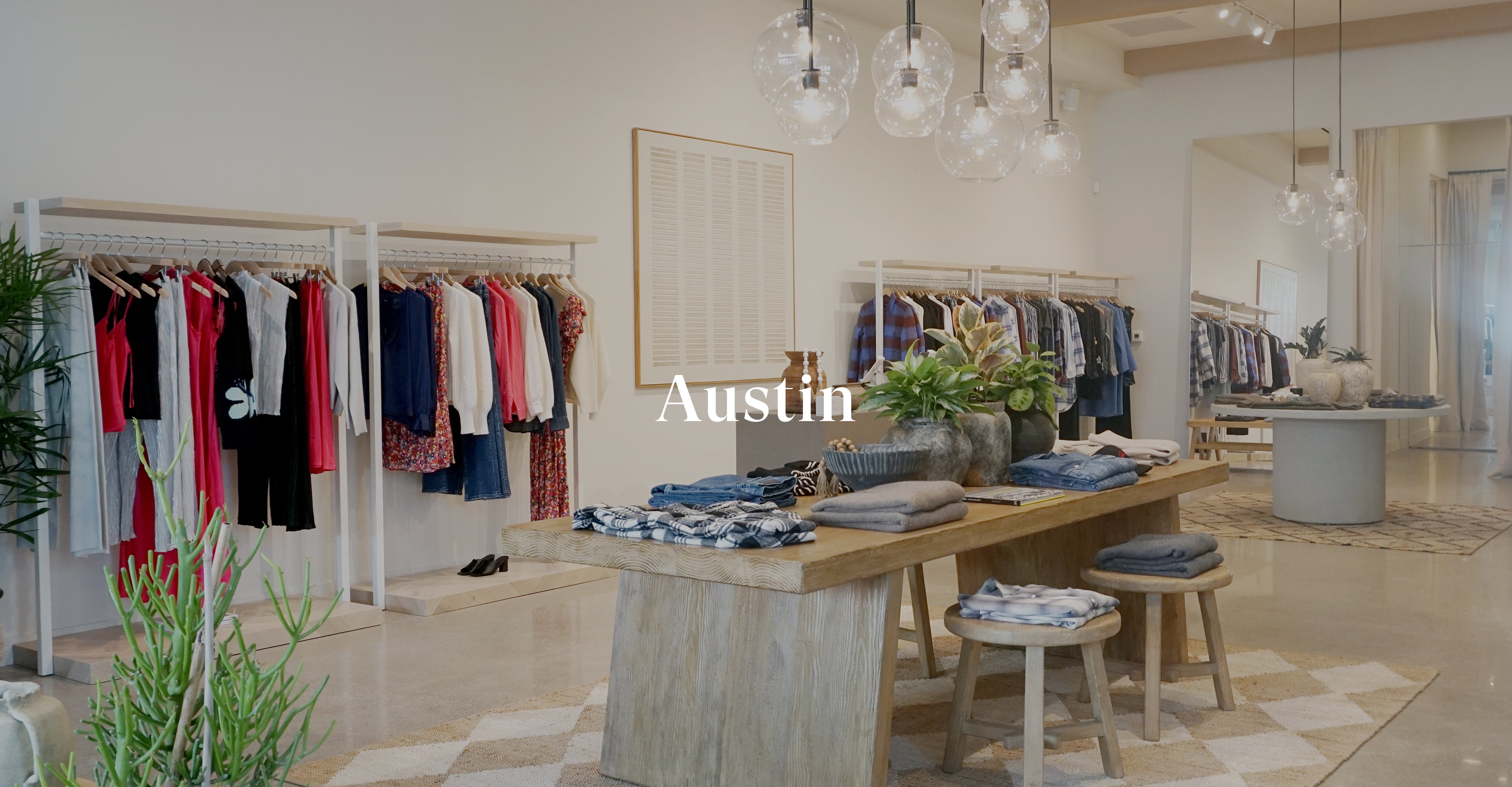 INSIDE OF AUSTIN STORE SHOWING CLOTHING RACKS OF CLOTHING AGAINST THE BACK WALL AND A LARGE TABLE WITH CLOTHING FOLDED ON TOP OF IT IN THE CENTER