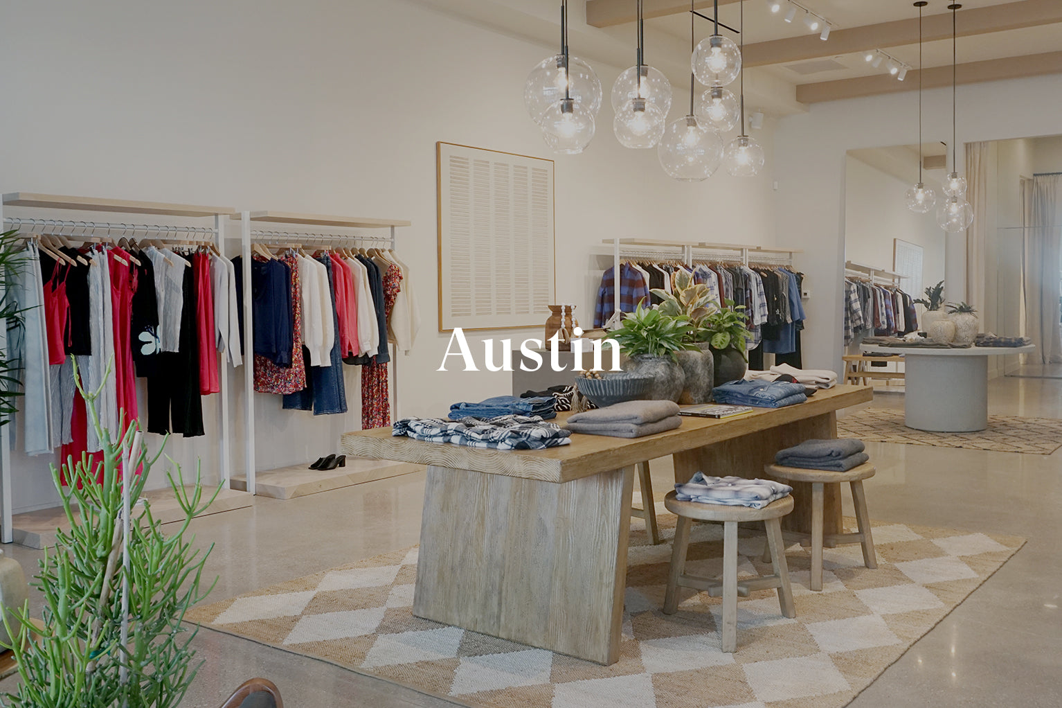 INSIDE OF AUSTIN STORE SHOWING CLOTHING RACKS OF CLOTHING AGAINST THE BACK WALL AND A LARGE TABLE WITH CLOTHING FOLDED ON TOP OF IT IN THE CENTER