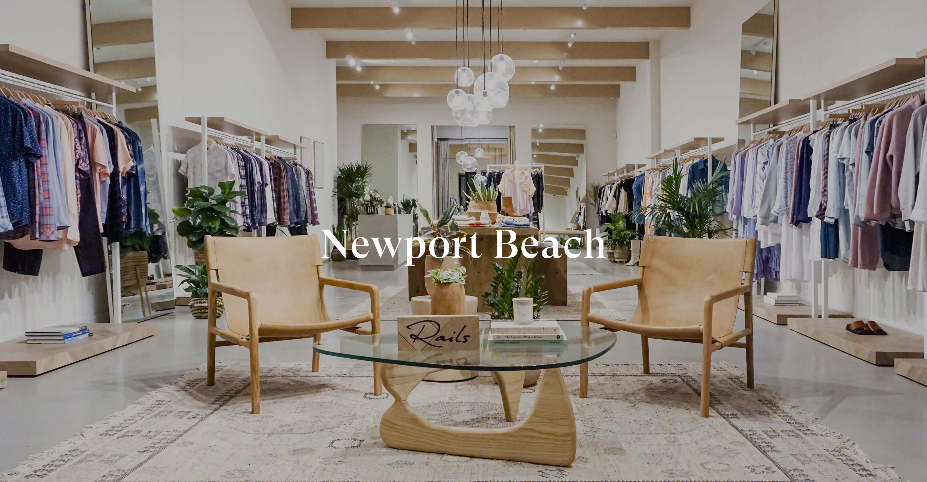 INSIDE OF NEWPORT BEACH STORE SHOWING MULTIPLE CLOTHING RACKS FULL OF CLOTHES AGAINST EACH WALL AND TWO CHAIRS IN THE MIDDLE OF THE ROOM
