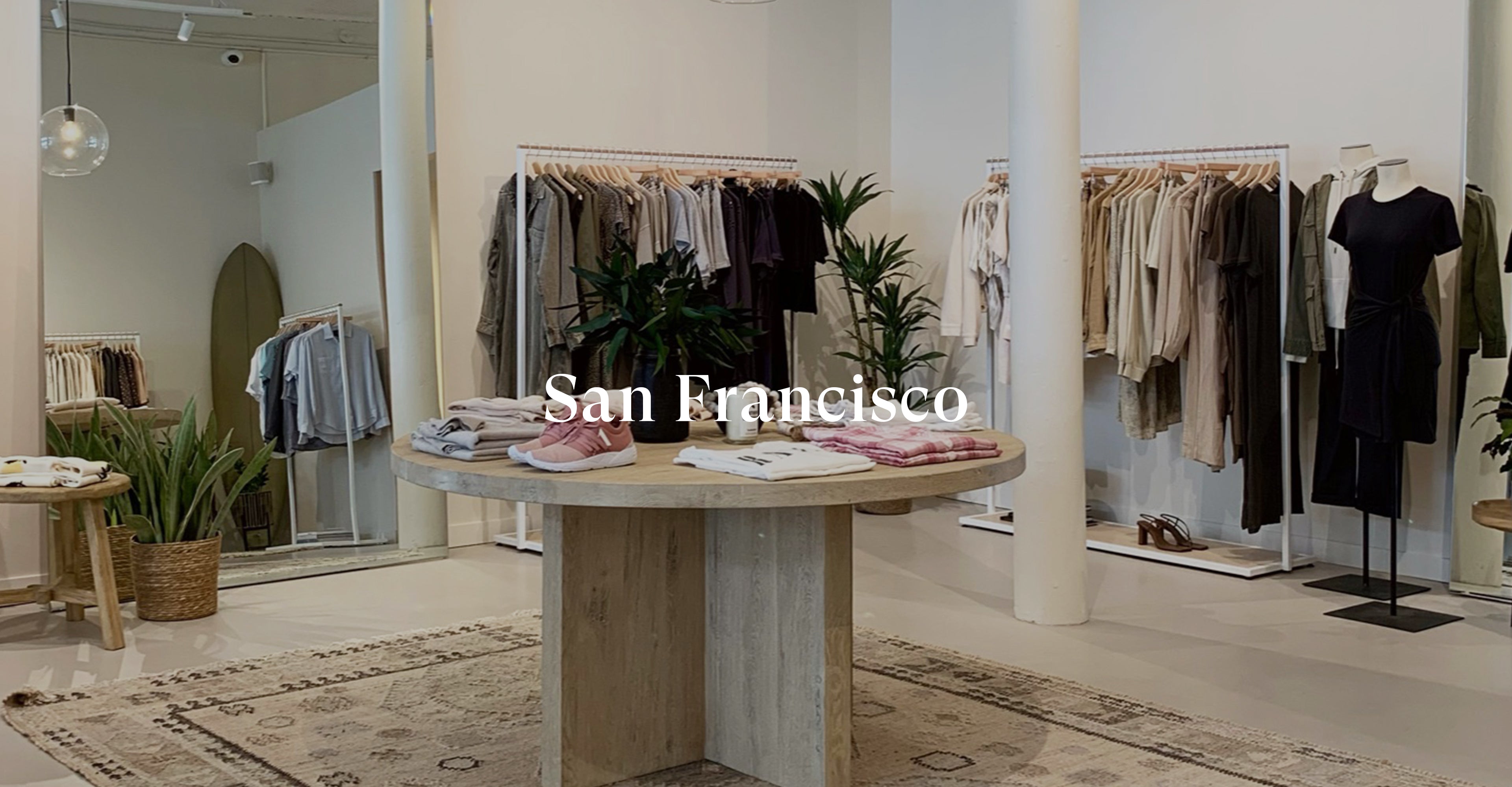 INSIDE OF SAN FRANCISCO STORE SHOWING TABLE IN MIDDLE OF ROOM WITH FOLDED CLOTHING ON IT AND MULTIPLE CLOTHING RACKS FULL OF CLOTHING AGAINST THE BACK WALL