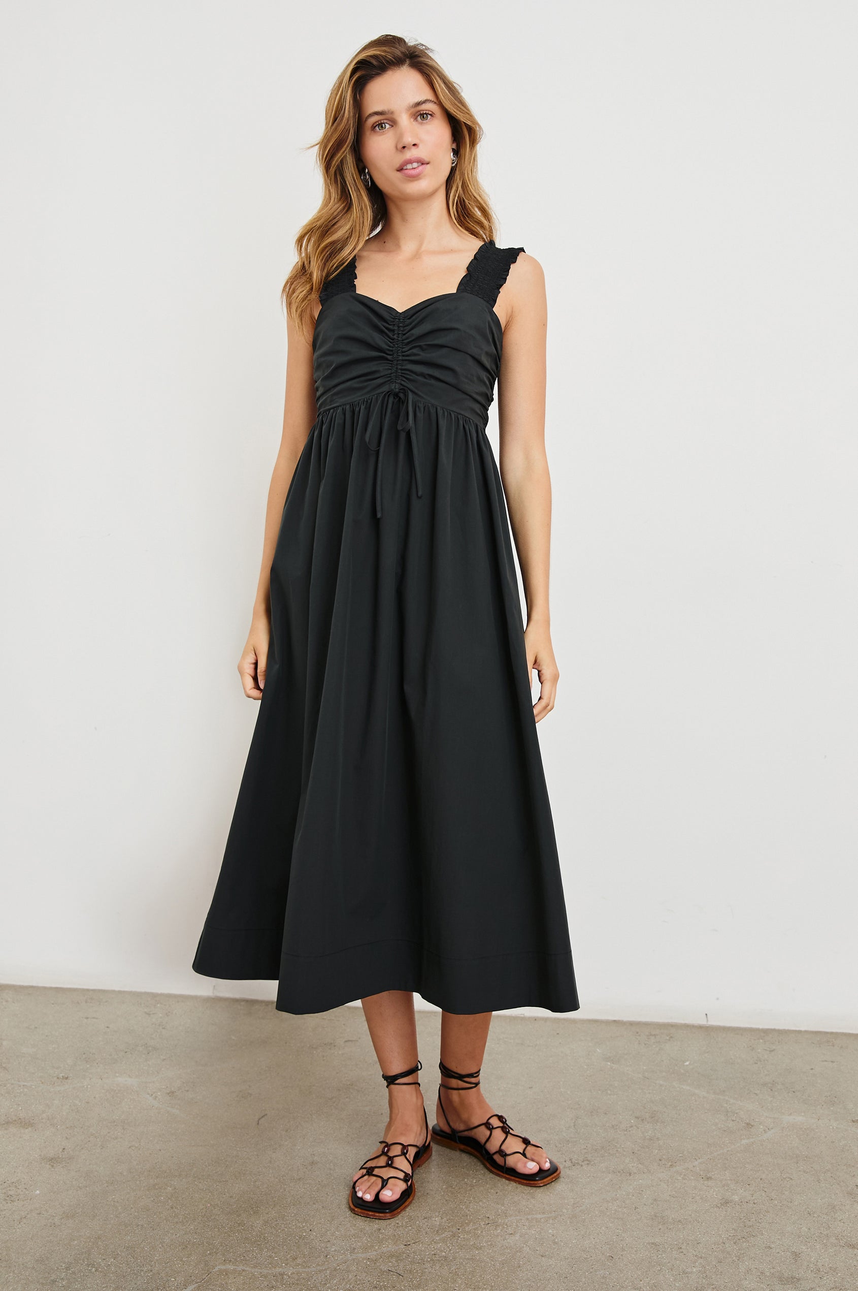 Irisa Sleeveless Midi Dress with Wide Straps, a Sweetheart Neckline, Horizontal Pleats on the Bodice, and a Tie and Vertical Pleats at the Waistline in Black Colorway - Front Full Body View Featured on Model 