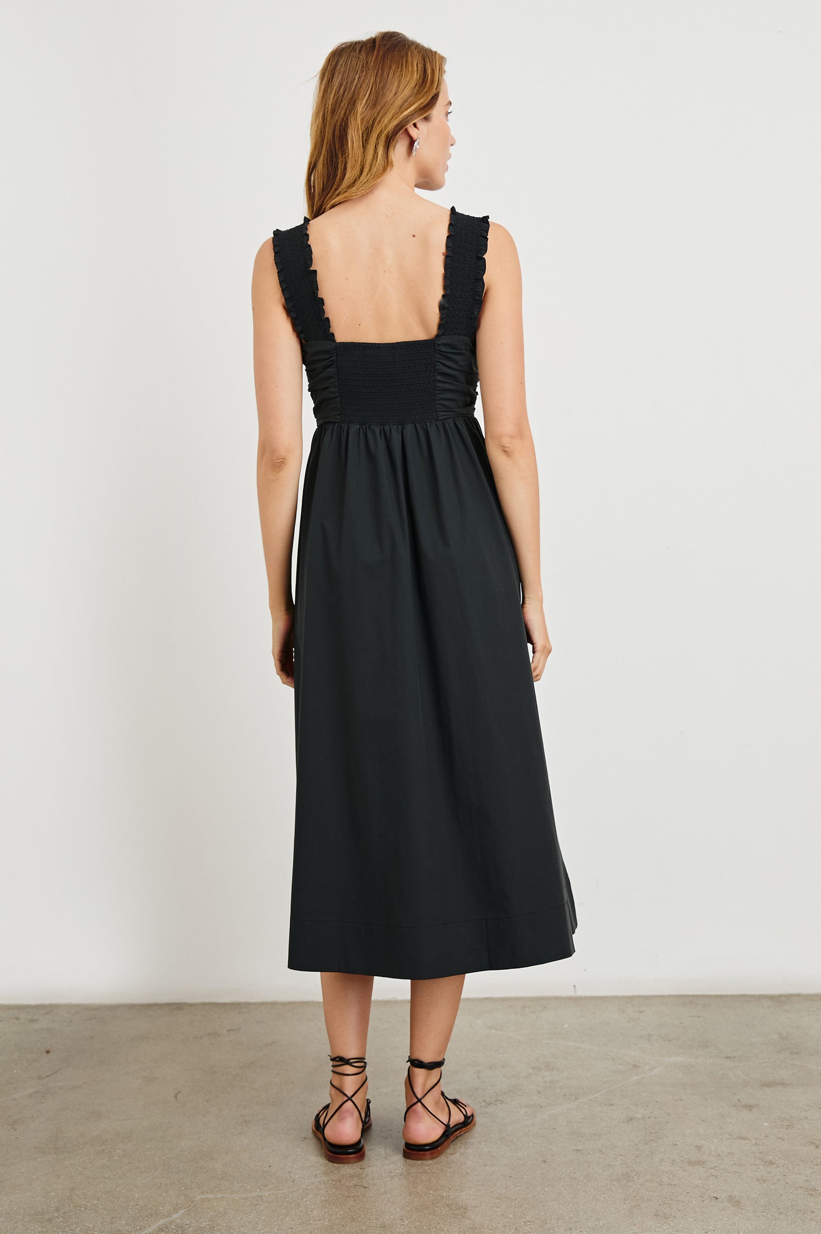 Irisa Sleeveless Midi Dress with Wide Straps, a Sweetheart Neckline, Horizontal Pleats on the Bodice, and a Tie and Vertical Pleats at the Waistline in Black Colorway - Back Full Body View Featured on Model 