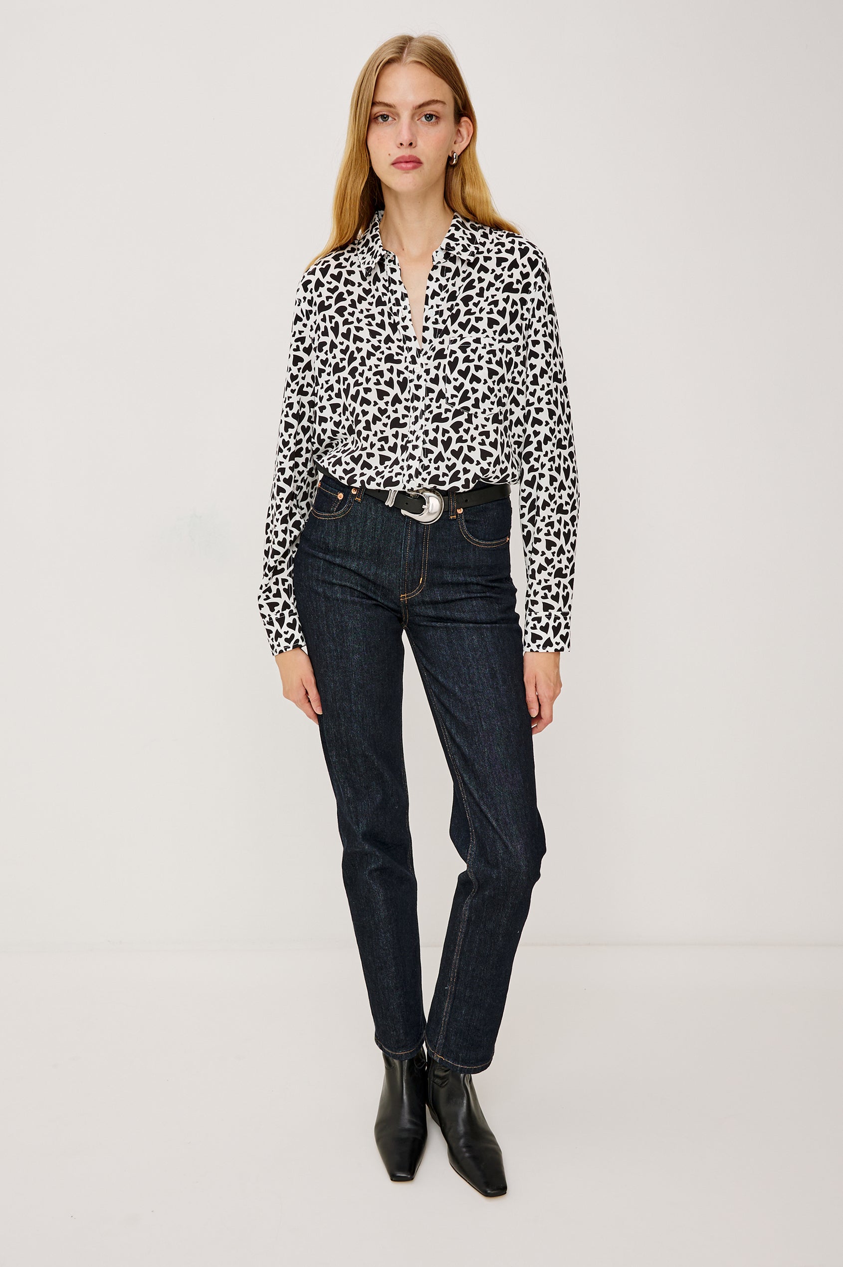 A white, long-sleeved button-down shirt featuring an all-over print of small black hearts, styled with a classic collar and a single chest pocket - front full body view on model