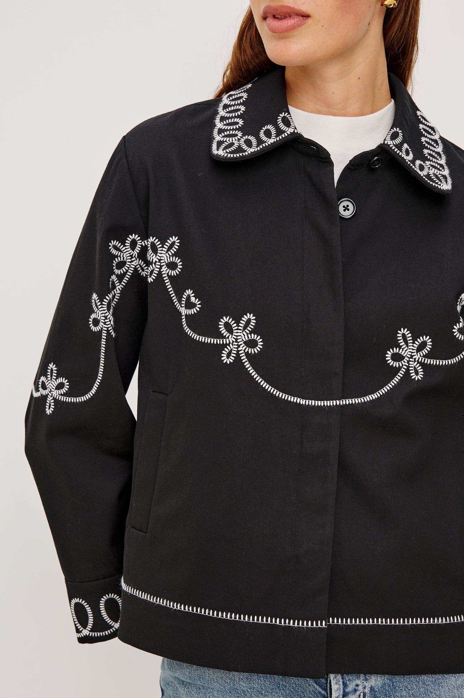 The Keaton is a collared button front jacket with contrast white western inspired embroidery on cuffs, collar, hem, sleeves and bodice in Black colorway. Front detail view featured on model.