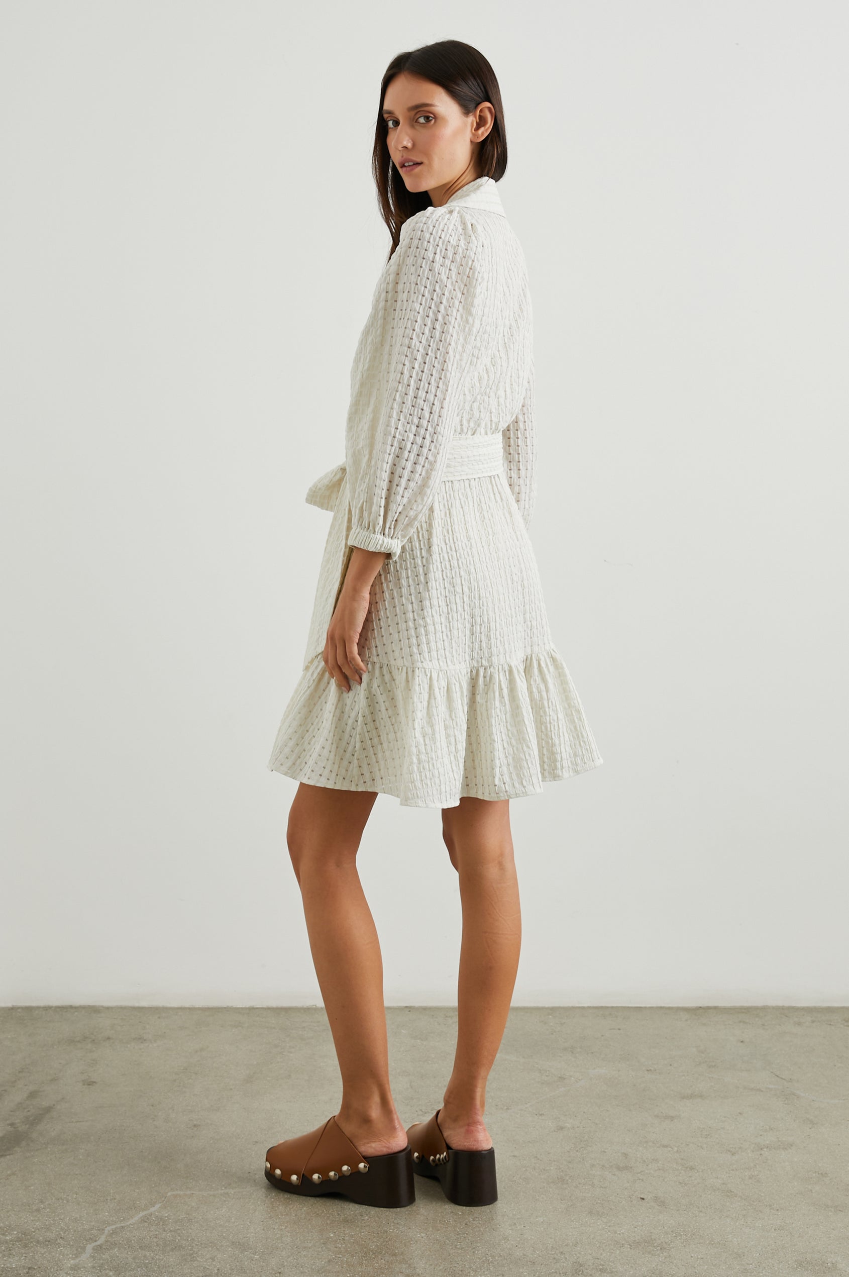 Kiara Mini Dress with Three-Quarter Sleeves, a Fabric Waist Tie, a Deep V Neck, a Collar, a Ruffled Hem and Textured Fabric in Ivory Seersucker Colorway - Side Full Body View Featured on Model 