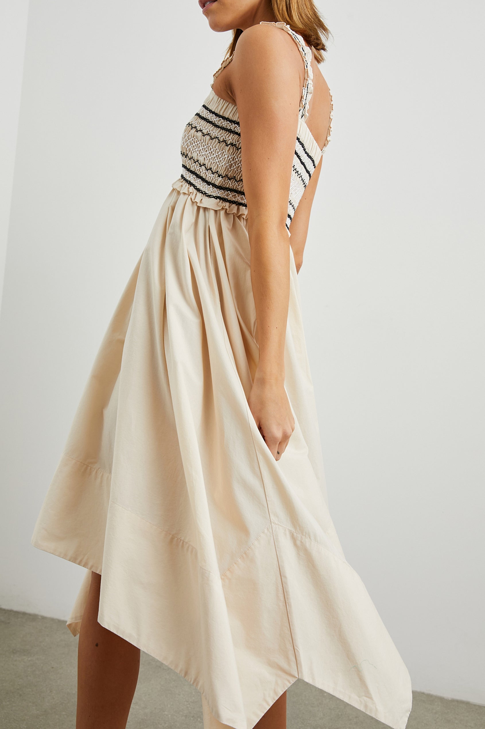 Kirsten Sleeveless Midi Dress with an Asymmetrical Hem, a Square Neckline, Shoulder Straps, a Shirred Bodice, Ruffled Trim on the Bodice, Black and White Stitching Details on the Bodice in Flax Colorway - Side View Featured on Model 