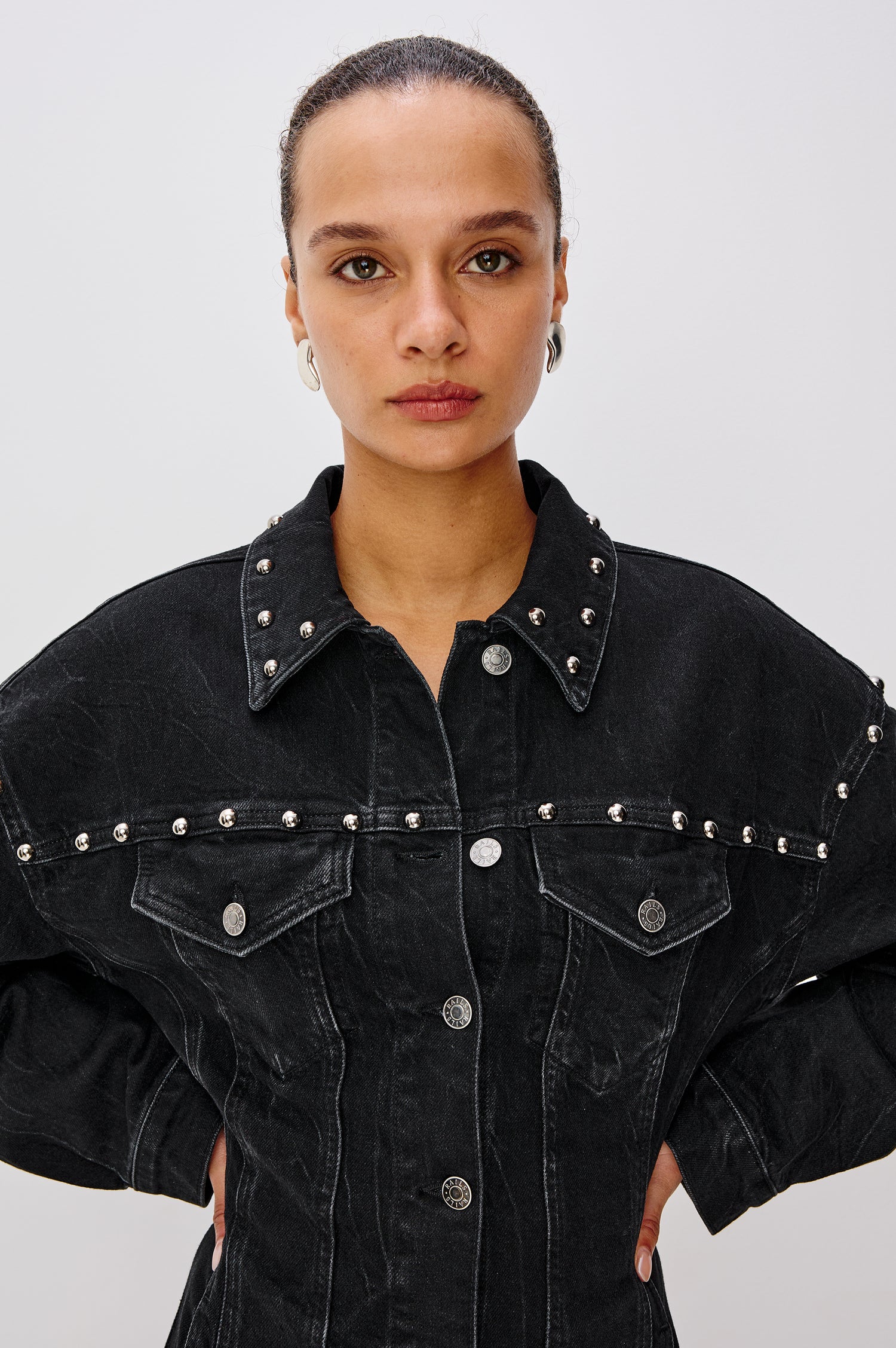 Ladera is a trucker style denim mini dress with long sleeves, drop shoulders, relaxed balloon sleeves, nail-back studs, and front button closures, with a subtle A-line shape and adjustable back strap in Black Stone Studded colorway. Front detail view featured on model.