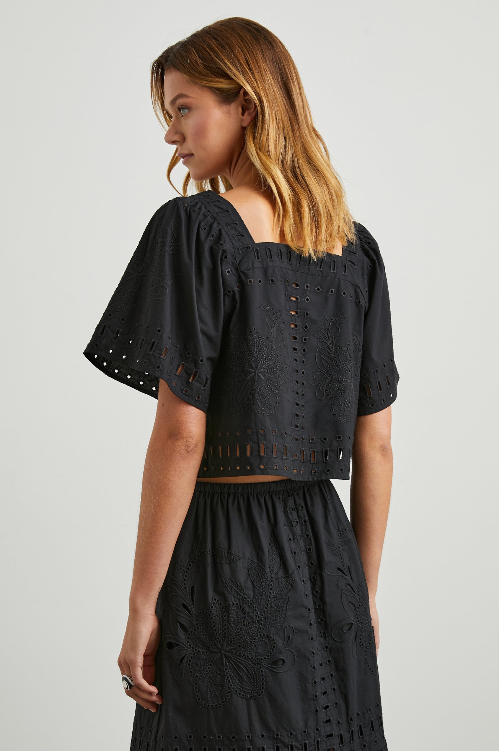 Laine Short Sleeve Crop Top with Square Neckline, and a Floral and Geometric Eyelet Design in Black Colorway - Back View Featured on Model