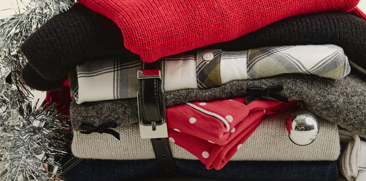 A stacked pile of folded winter clothing and accessories, including a bright red knit top, black knit layers, plaid shirts, gray wool, a red polka-dot scarf, and a silver metallic ornament, with silver tinsel on the left side.