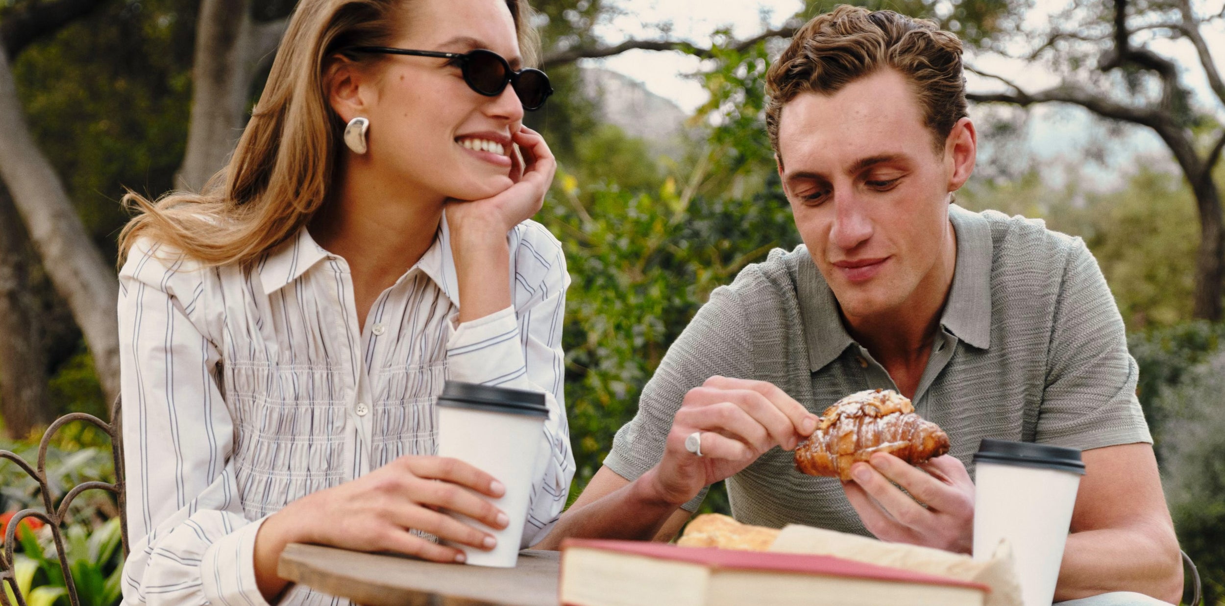 A man and a woman sitting at an outdoor table, with the man holding a pastry and the woman holding a takeaway coffee cup. They are wearing casual clothing, and the background shows blurred trees and greenery.