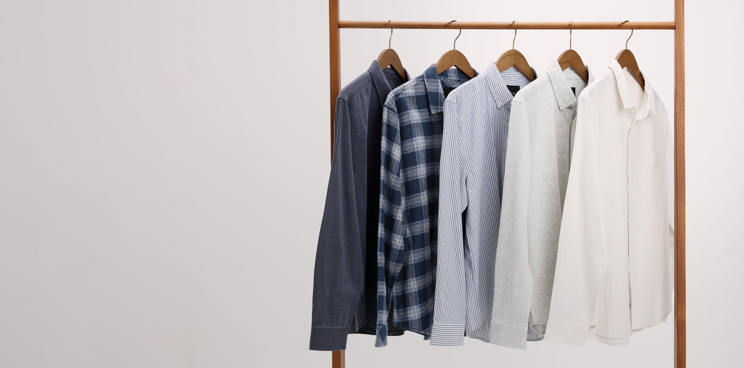 Five long-sleeve button-up shirts in different colors and patterns hang on a wooden clothing rack against a plain white wall. The shirts include dark navy, blue plaid, light blue, light gray, and off-white.