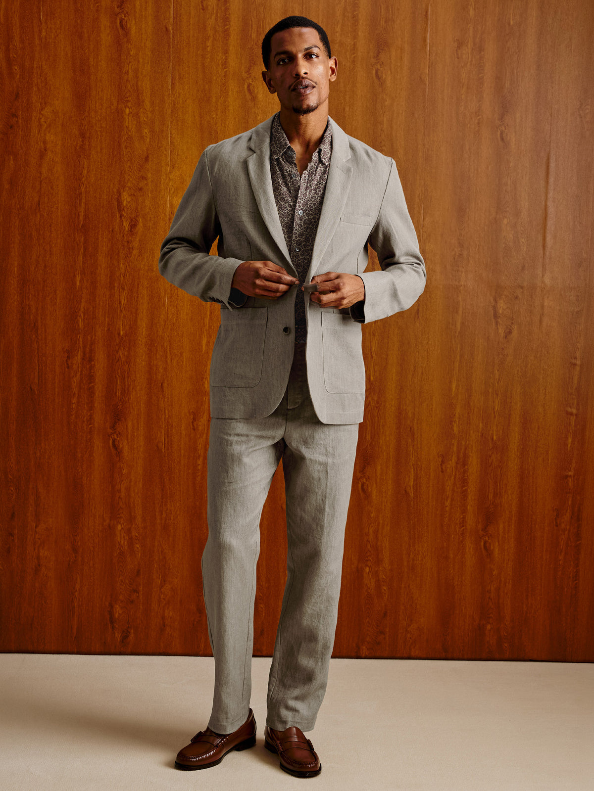 A man wearing a light gray two-piece suit with a patterned shirt and brown leather loafers, standing against a warm wood-paneled wall.