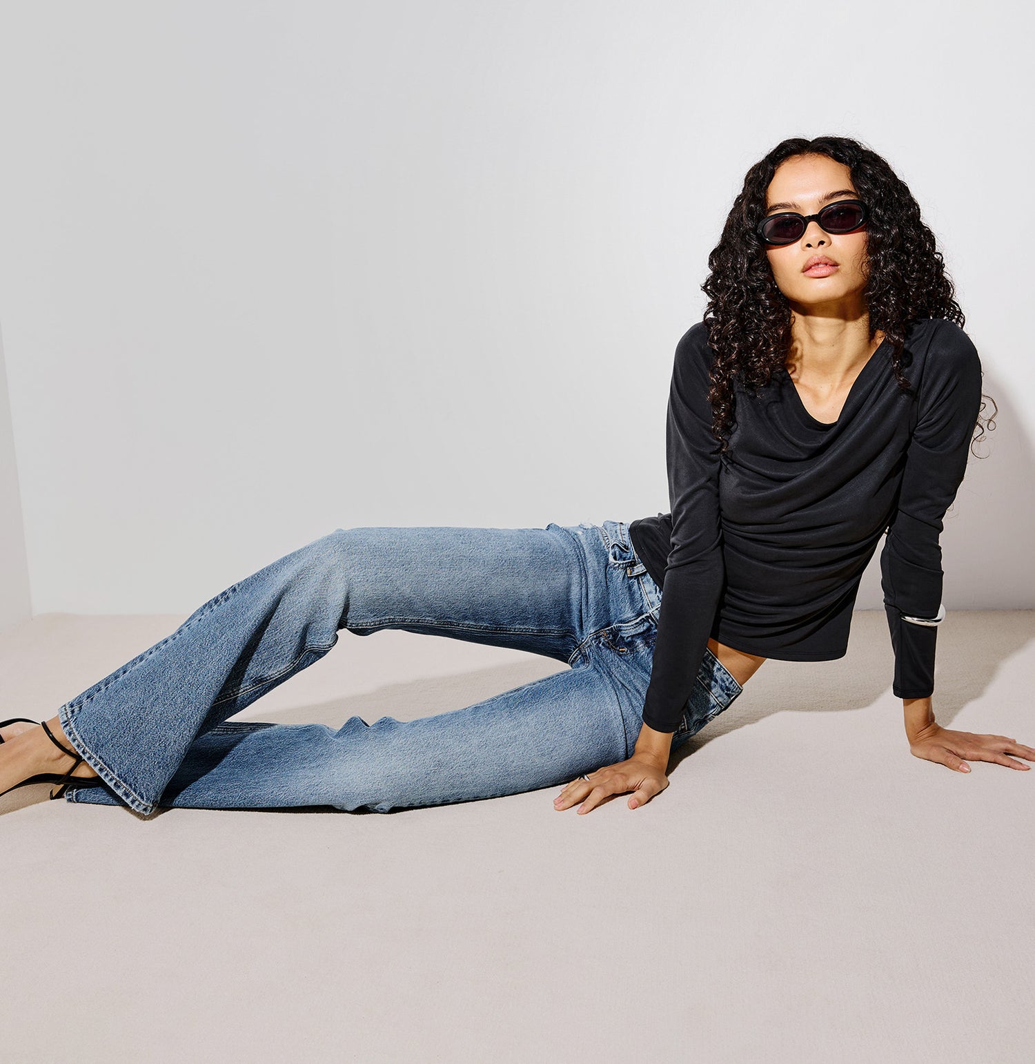 A model wearing a black long-sleeve cowl-neck top and light-wash blue flare jeans, posing on a light beige studio floor against a white background.