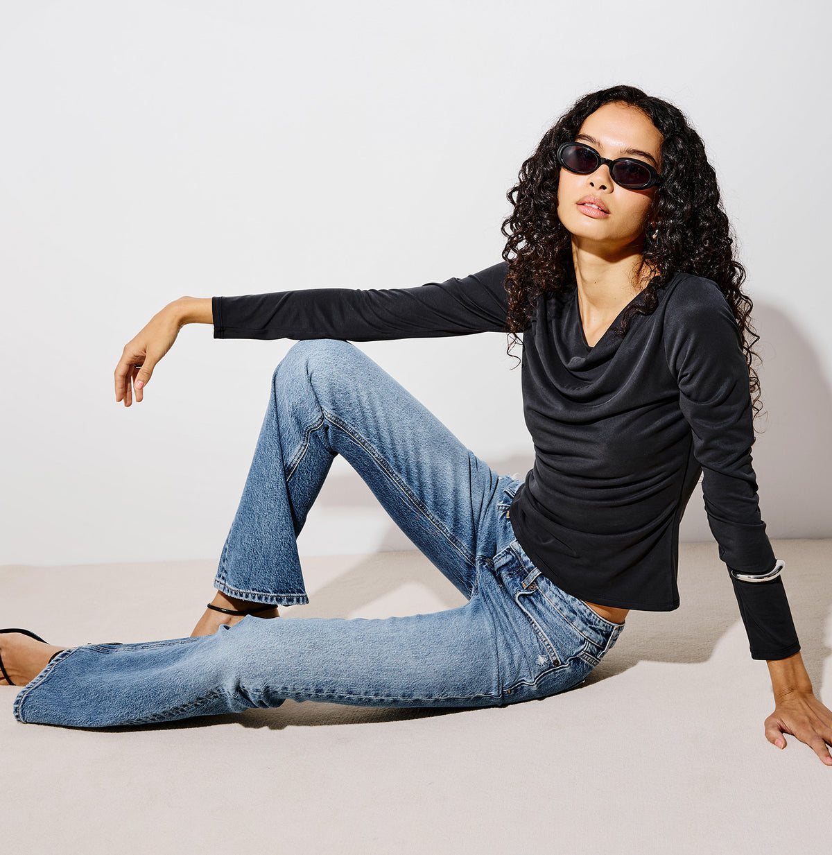 A woman wearing a black long-sleeve cowl-neck top, blue flare jeans, and black strappy heels sits on the floor of a minimalist white studio corner.