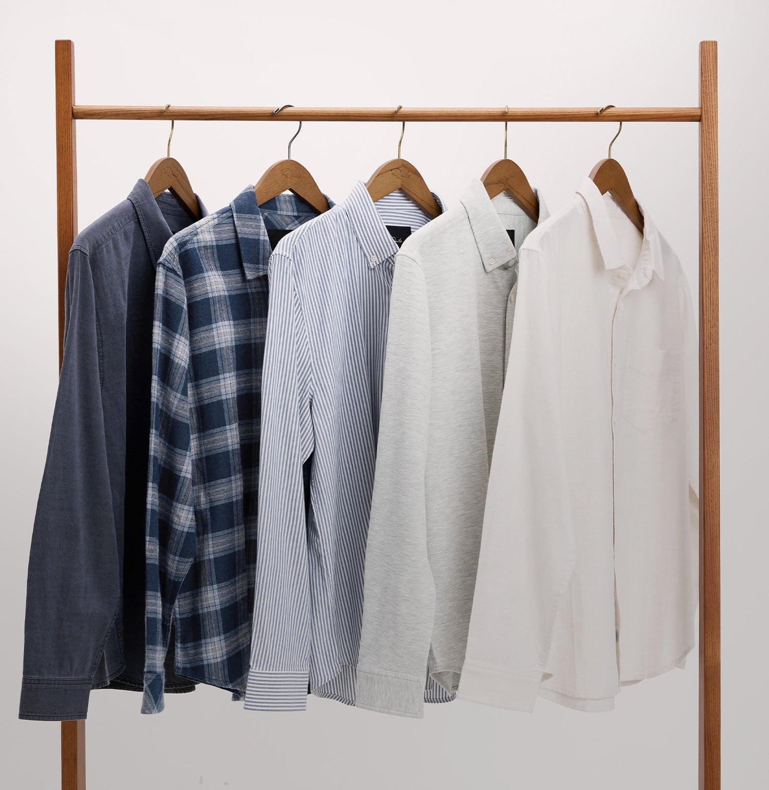 Six long-sleeve button-up shirts in various colors and patterns hang on wooden hangers from a wooden clothing rack against a plain light background.