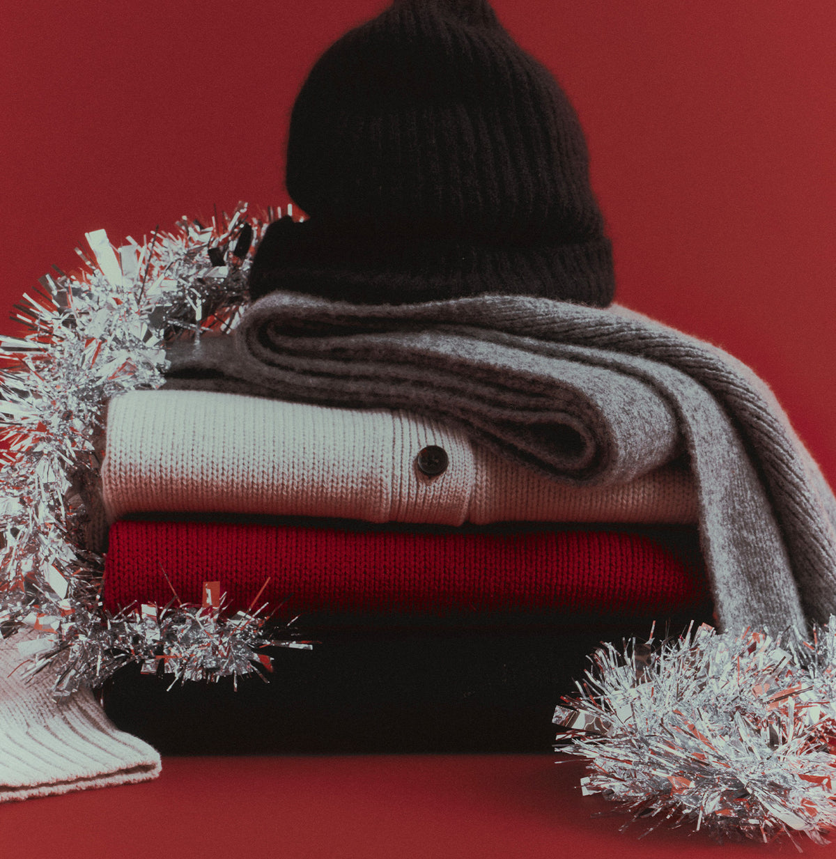 A neatly folded stack of knitwear, including a cream cardigan, a red sweater, and a gray scarf topped with a dark beanie, framed by silver tinsel against a red background.