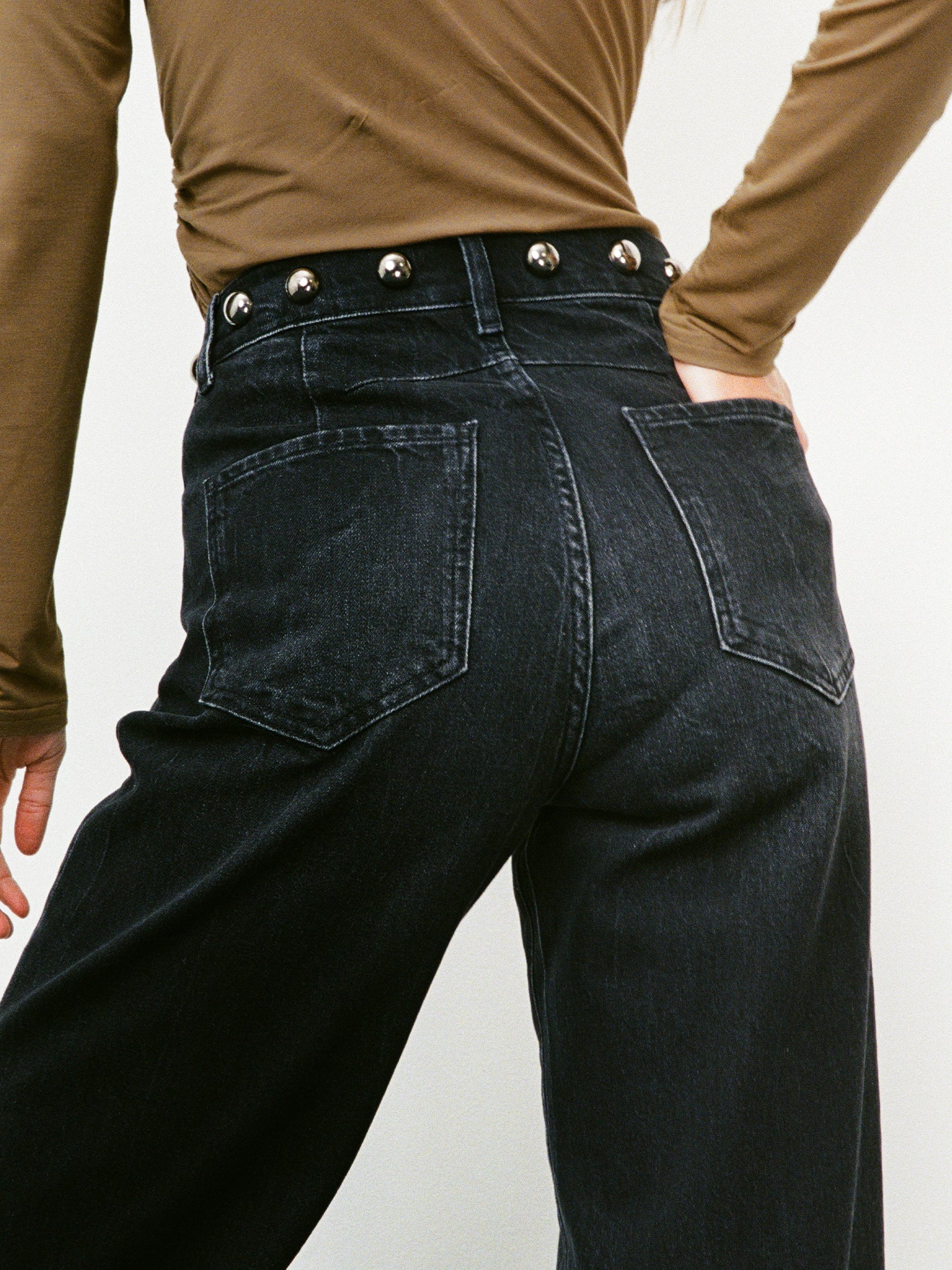 Woman wearing black wide-leg jeans with silver buttons on a plain background. 