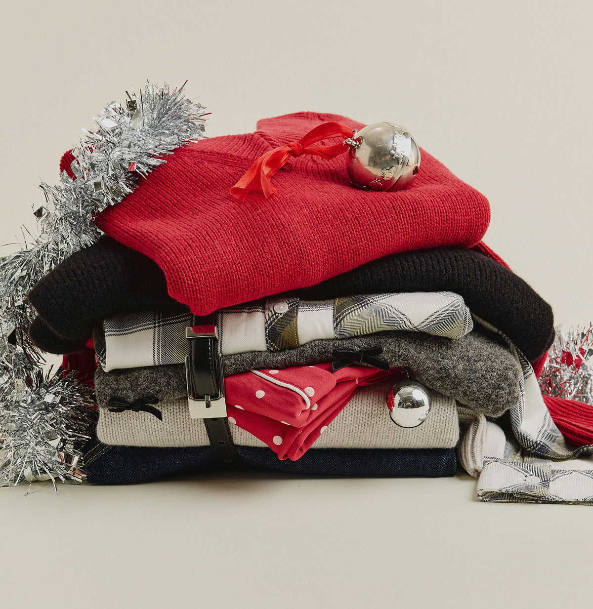 A styled stack of folded knit garments and accessories on a light beige background, including a bright red sweater, a black knit piece, gray plaid and heathered textiles, a red polka-dot scarf, and silver tinsel with a metallic ornament.