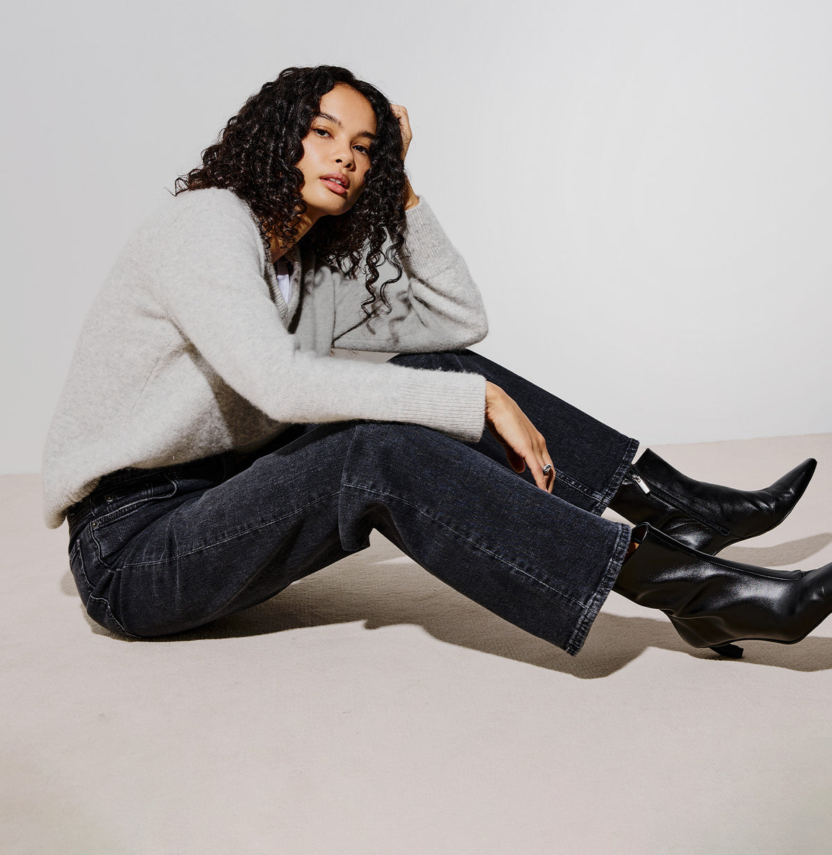 A woman sits cross-legged on a light beige floor against a white studio wall, wearing a light gray knit sweater, dark wide-leg jeans, and black ankle boots.