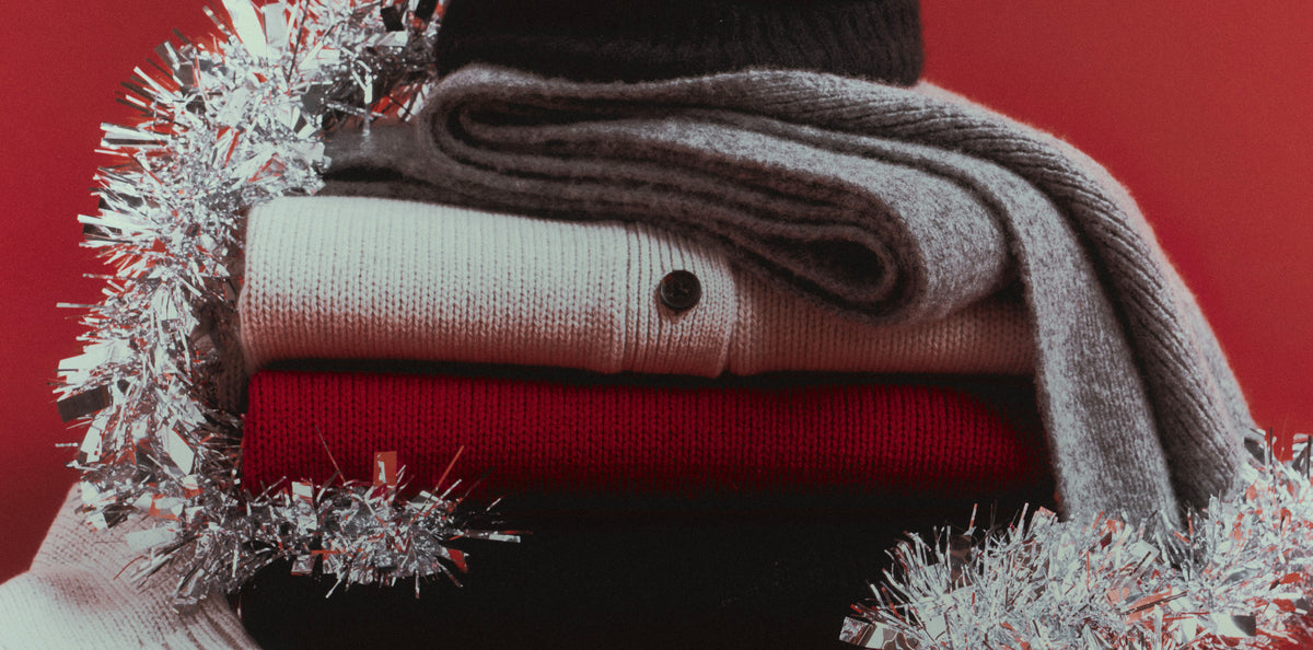 A stack of folded knit garments in gray, cream, and red, styled with silver tinsel against a solid red background.