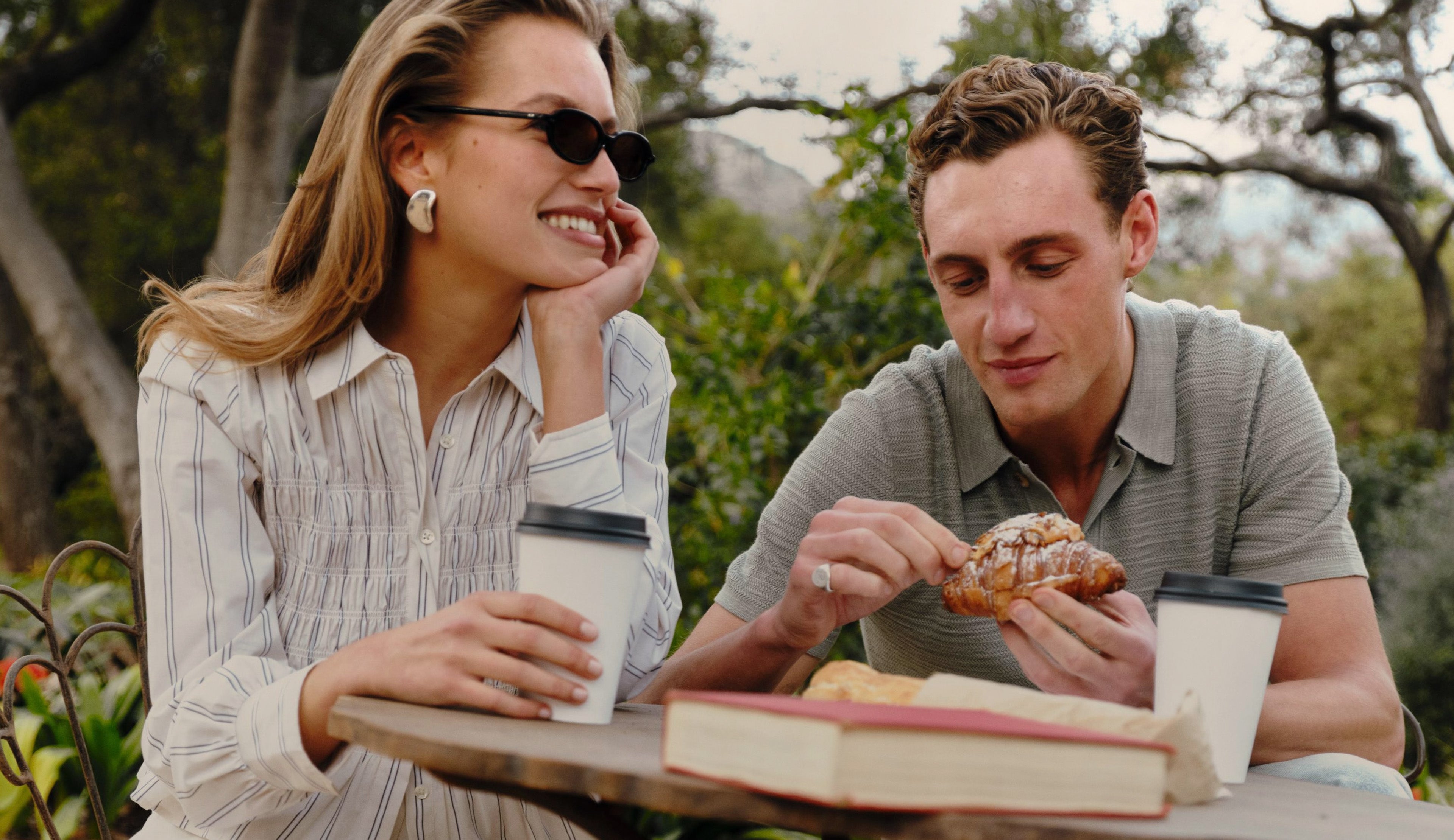 A man and a woman sitting at an outdoor table, smiling and laughing while having coffee and eating pastries. The woman is wearing black sunglasses and a white striped button-up shirt, and the man is wearing a gray polo shirt.