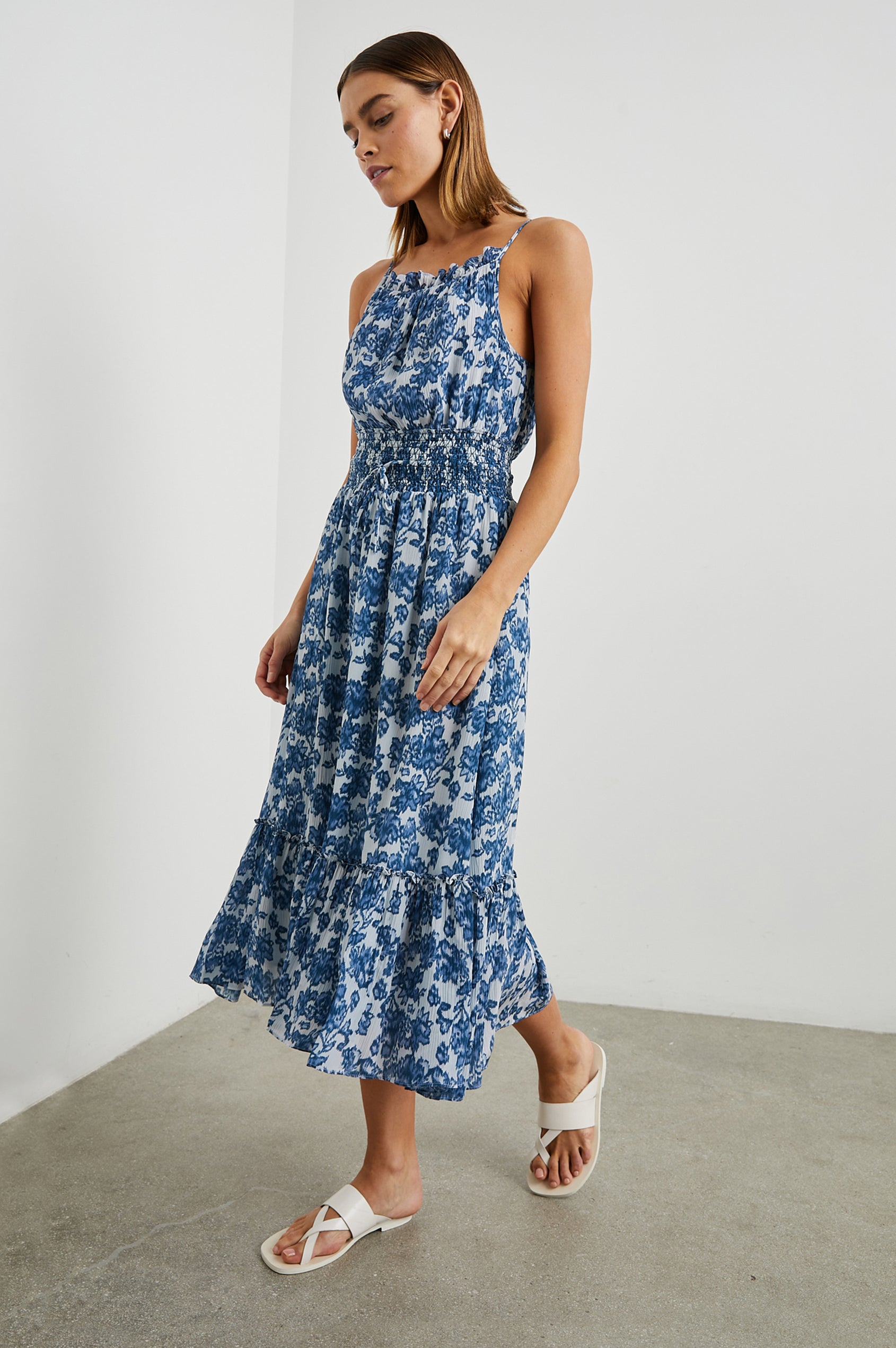Magdalene Sleeveless Midi Dress with Spaghetti Straps, a Button Keyhole at the Back, a Shirred Waist with a Drawstring, a Scoop Neckline with Ruffled Trim, and a Ruffled Hem in Chambray Floral Colorway, Blue Flowers on White - Side Full Body View Featured on Model  