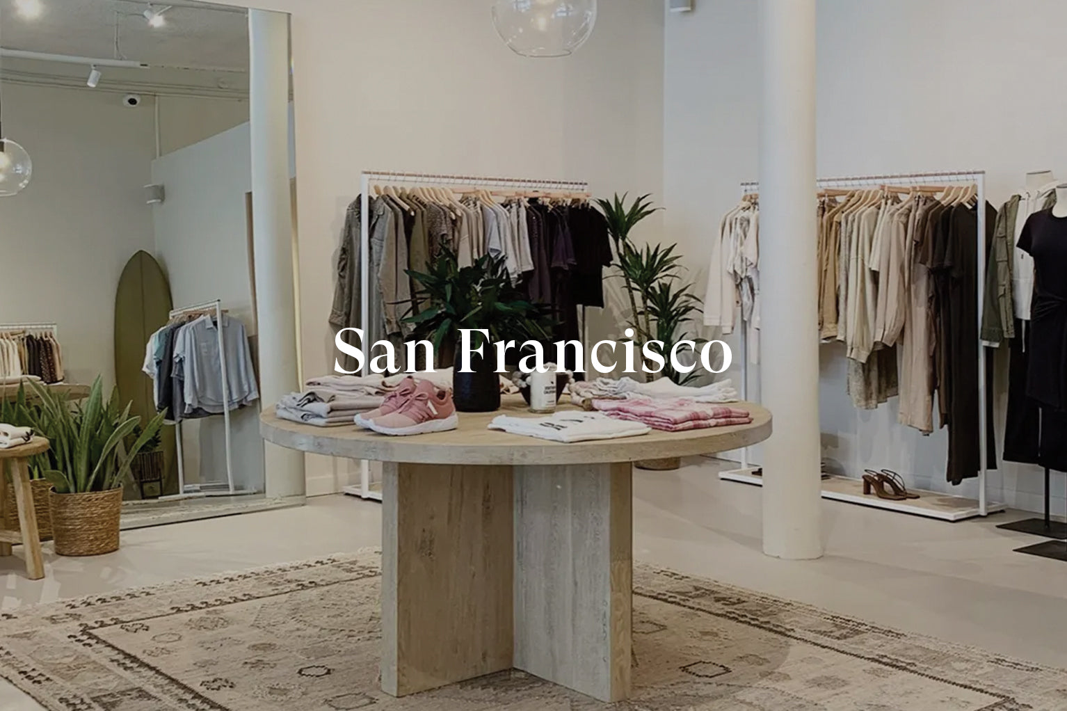 INSIDE OF SAN FRANCISCO STORE SHOWING TABLE IN MIDDLE OF ROOM WITH FOLDED CLOTHING ON IT AND MULTIPLE CLOTHING RACKS FULL OF CLOTHING AGAINST THE BACK WALL