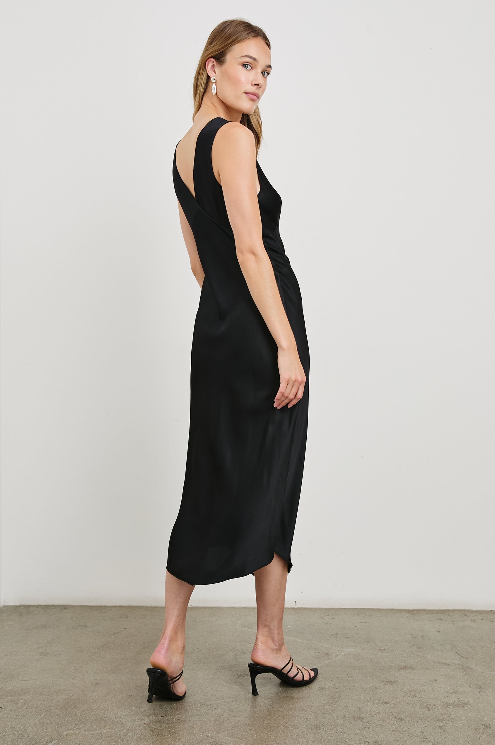 Monique Sleeveless Midi Dress with an Asymmetrical Hem Line, Side Slit, Deep V Neck, and Wide Shoulder Straps in Black Colorway - Back Full Body View Featured on Model