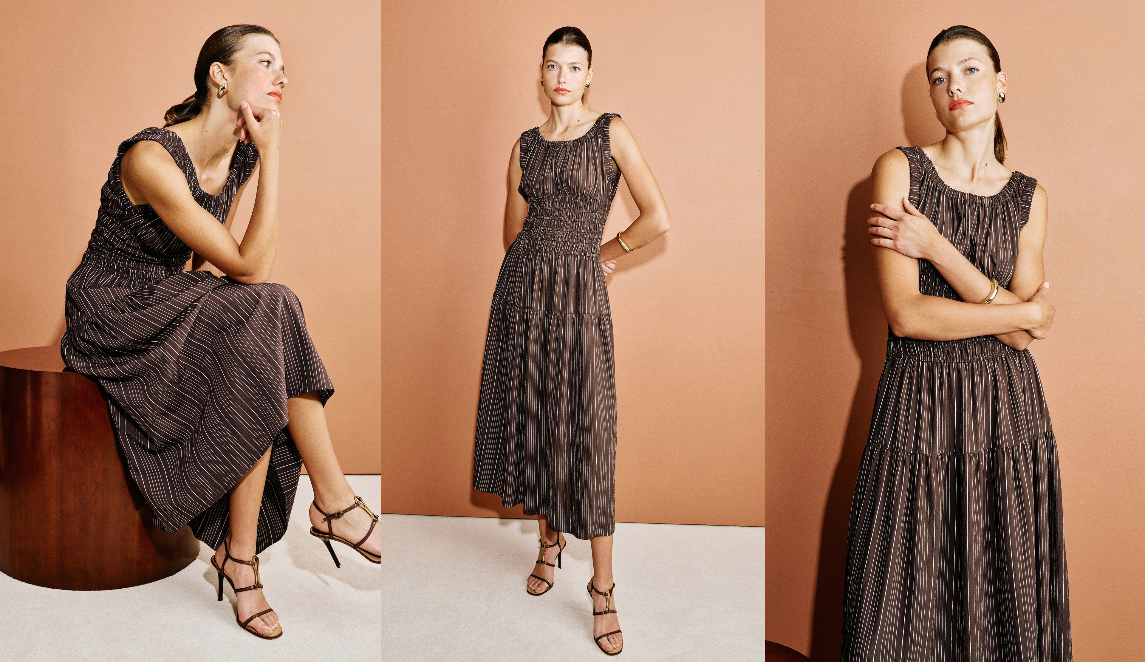A woman modeling a sleeveless pleated midi dress in a dark brown color, shown in three poses against a warm peach studio backdrop: seated on a wooden stool, standing with one hand on her hip, and standing with arms crossed.