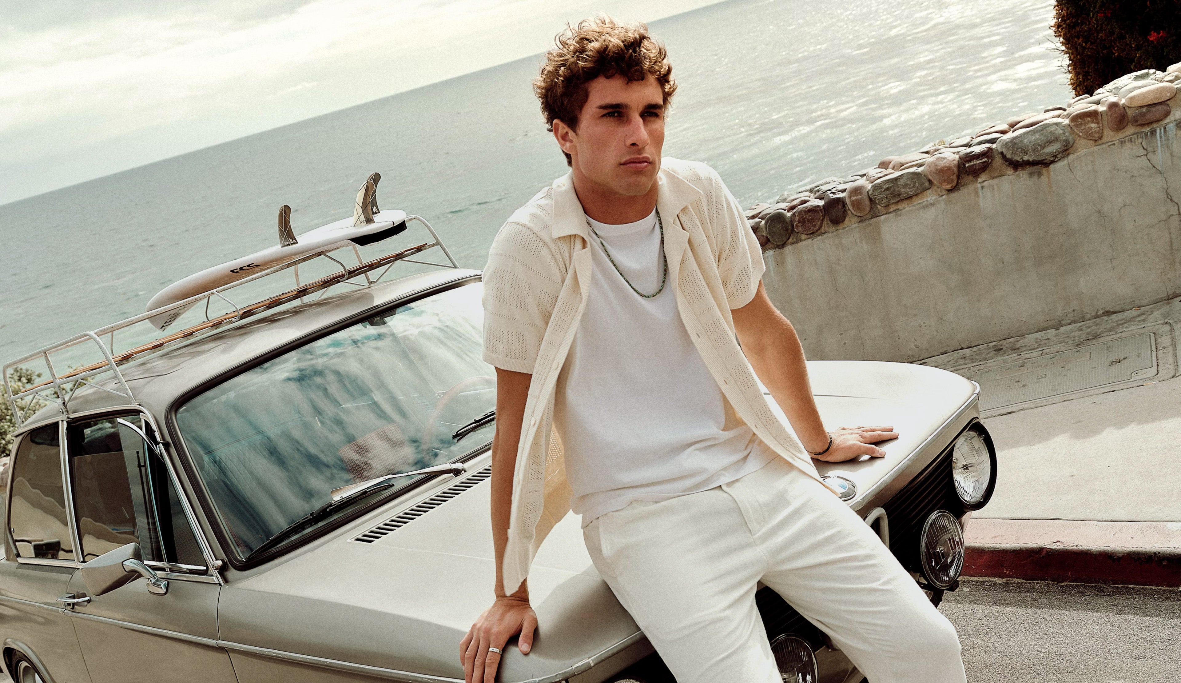 Man in white outfit leaning against a vintage car by the sea