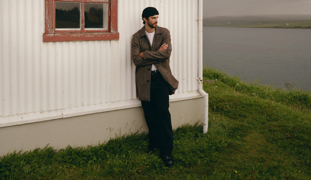 A man wearing a brown tweed jacket, black trousers, and a black beanie leans against the corner of a white house with a red-trimmed window, with grass and water in the background.