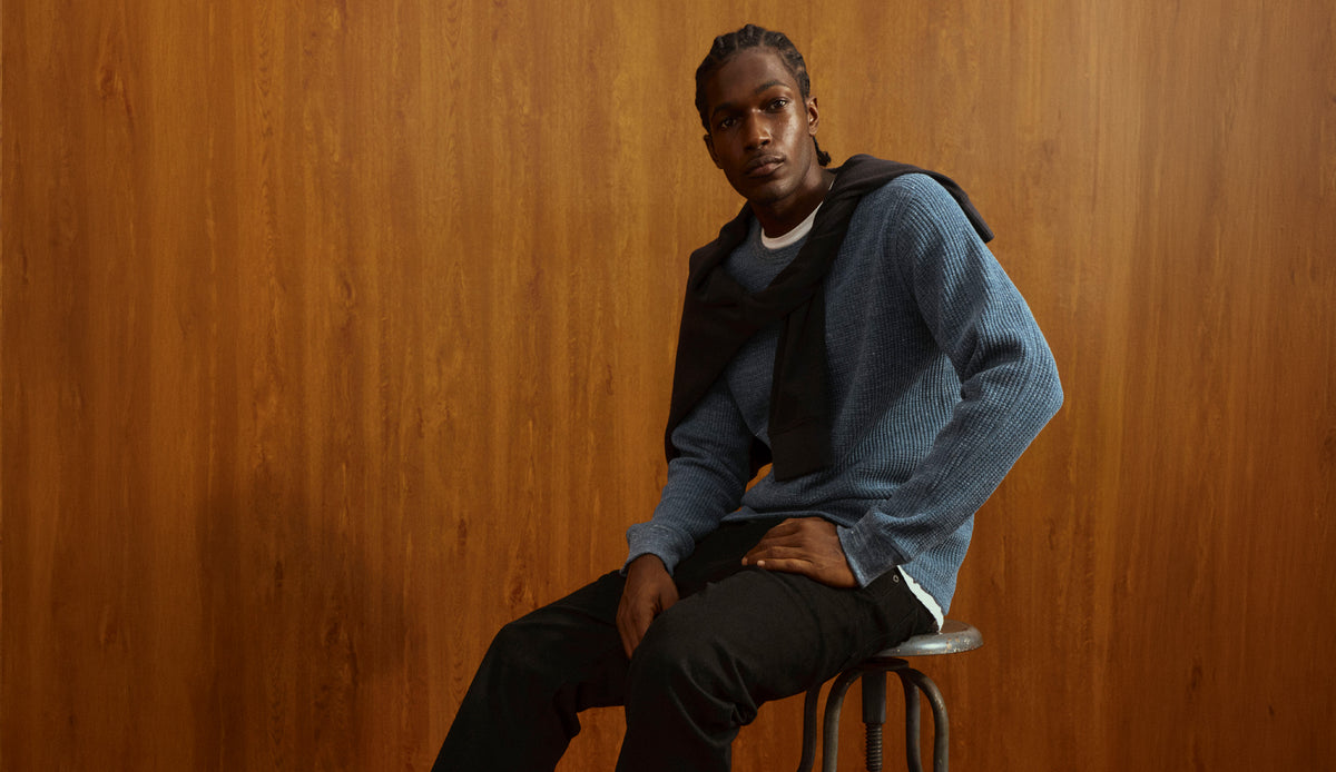 A man wearing a blue textured knit sweater, black hoodie, and black pants sits on a stool against a warm wood-paneled background.