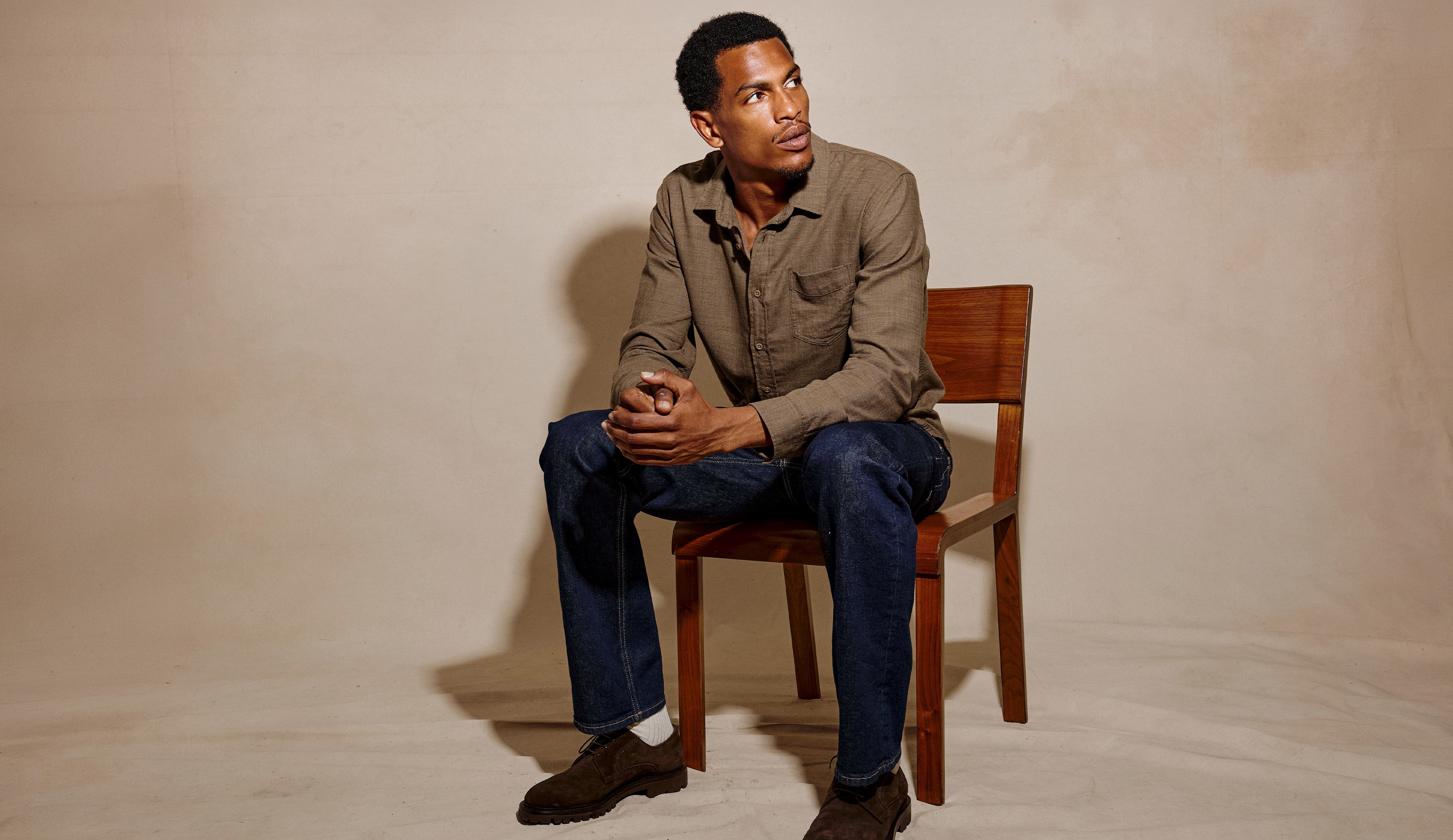 A man sits on a wooden chair wearing a brown button-up shirt, dark blue jeans, and brown shoes against a textured beige studio backdrop.