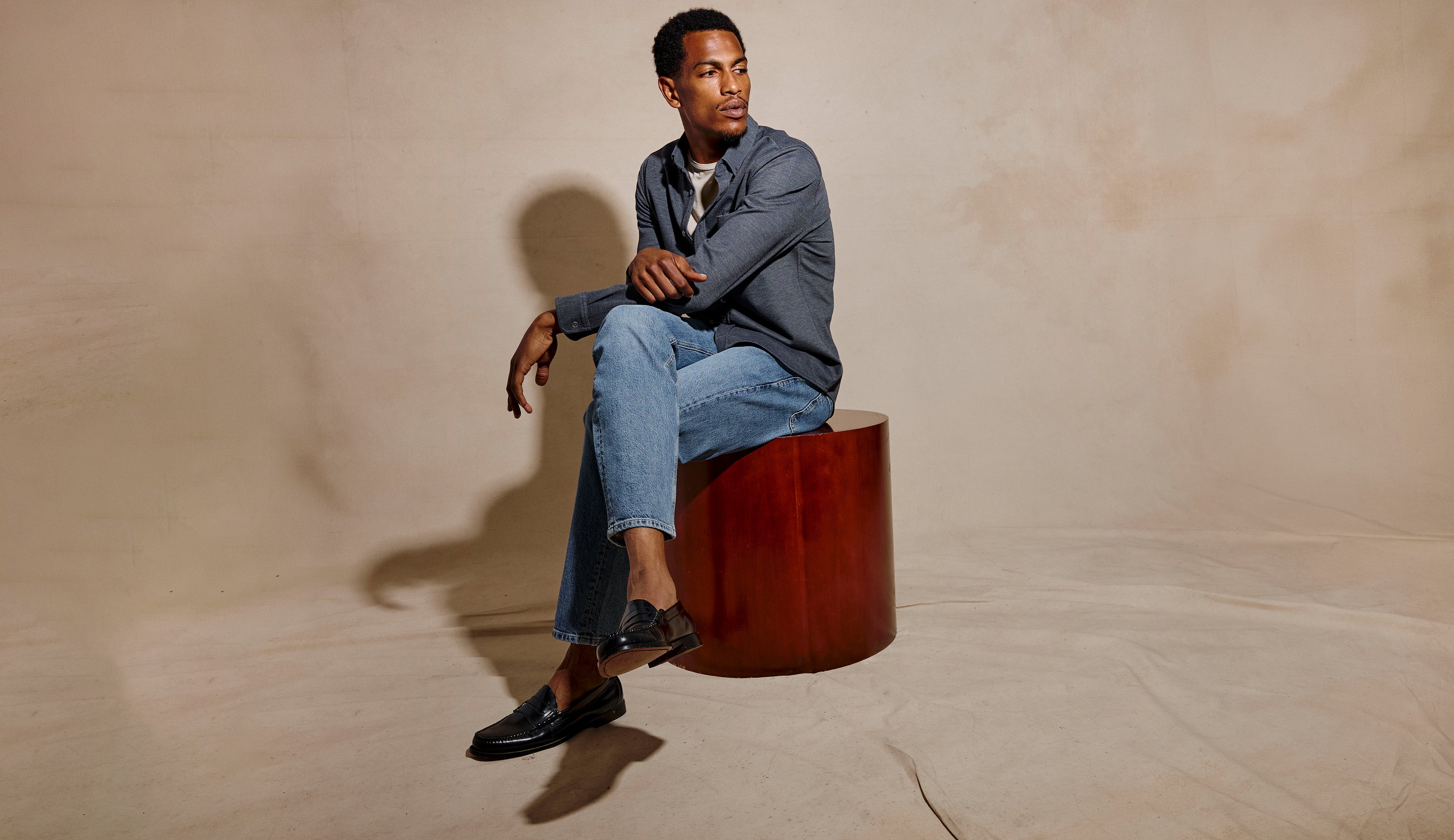 A man sits on a cylindrical wooden stool in a studio setting, wearing a gray blazer, light blue jeans, and black loafers against a warm beige backdrop.