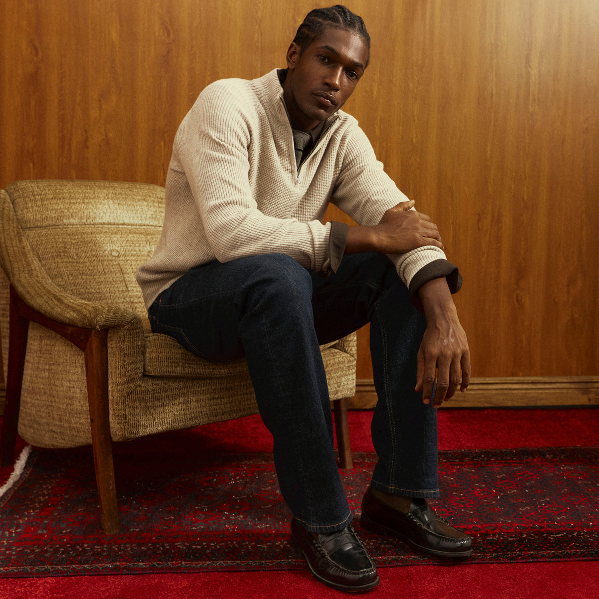 A man sits in a woven armchair against a wood-paneled wall, wearing a cream ribbed quarter-zip sweater, dark blue jeans, and black leather shoes on a red patterned rug.