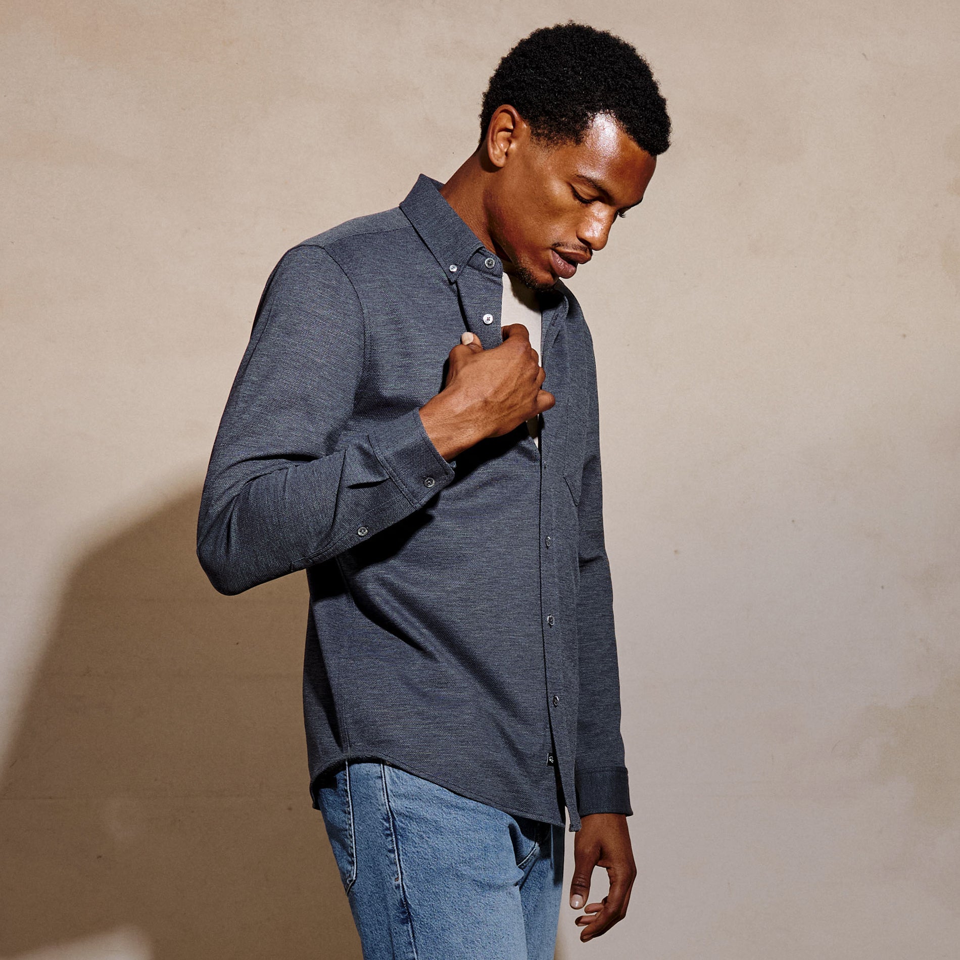 A man wearing a dark gray heathered long-sleeve button-down shirt and blue jeans, standing against a textured beige wall.